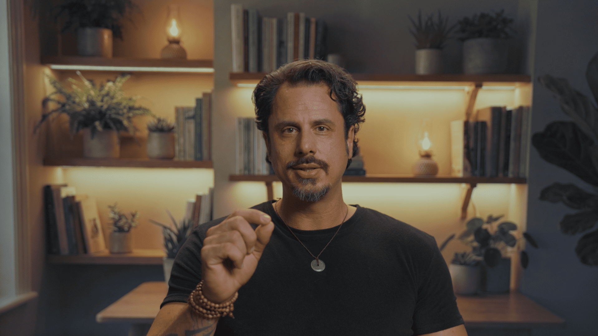 Man with short curly hair and beard gesturing with fingers in a warmly lit room with shelves and plants.