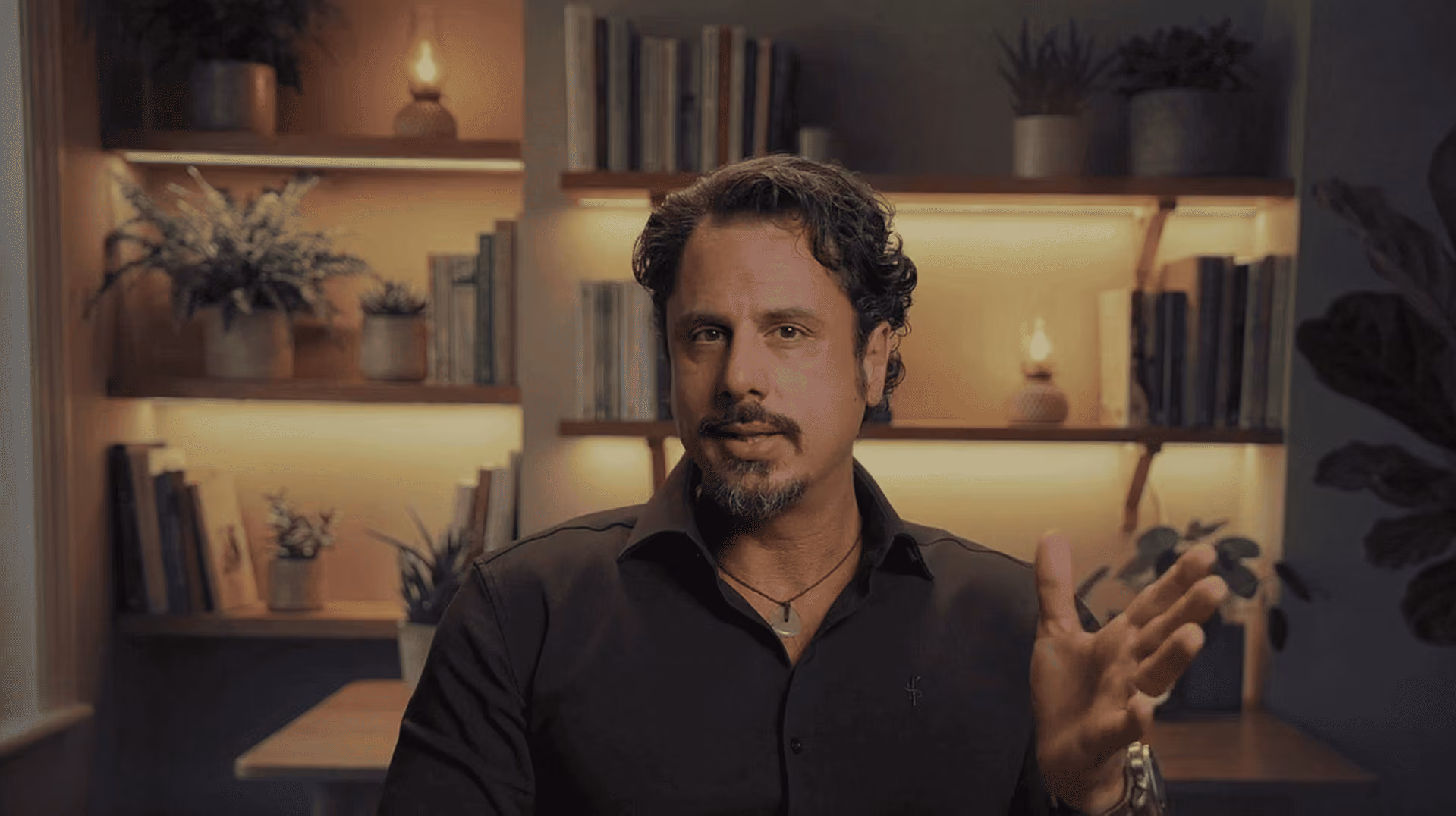 Man with dark curly hair and goatee gesturing with hand in a warmly lit room with books and plants.
