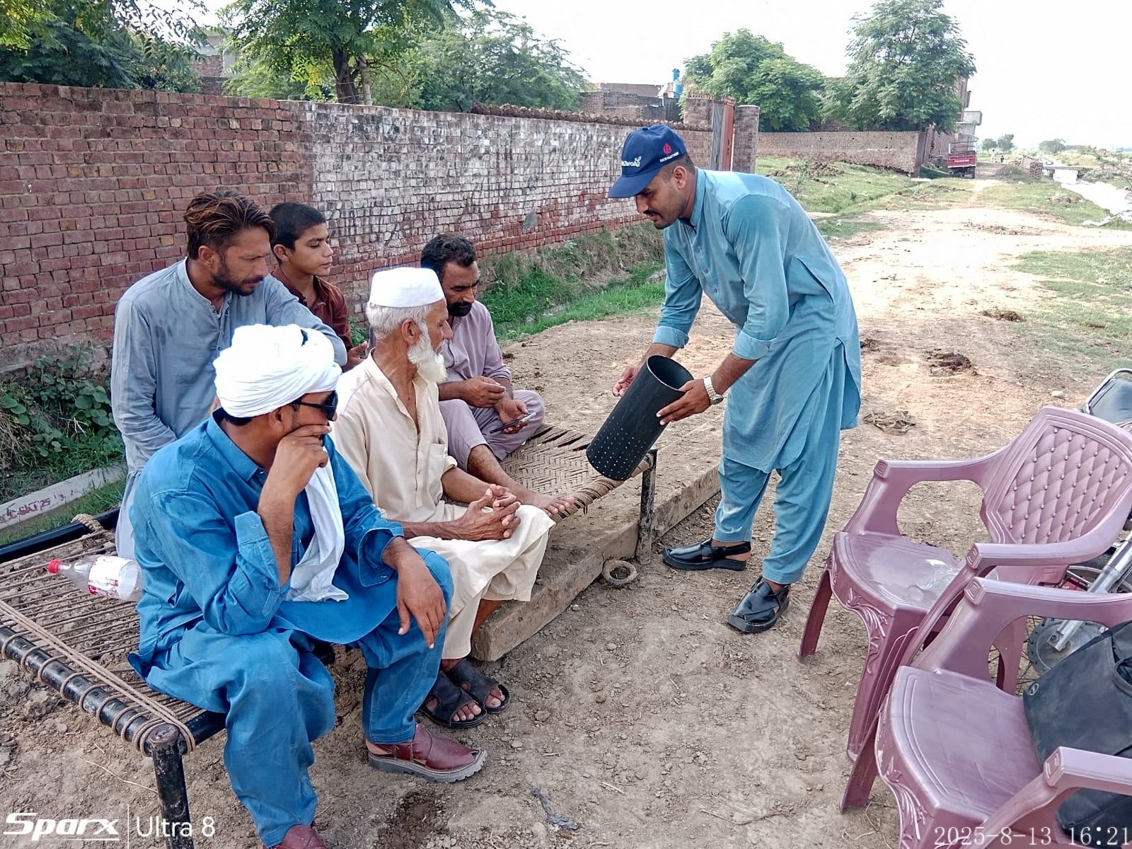 A group of men sitting outdoors on a traditional woven cot while one man in blue attire bends forward holding a perforated black container.