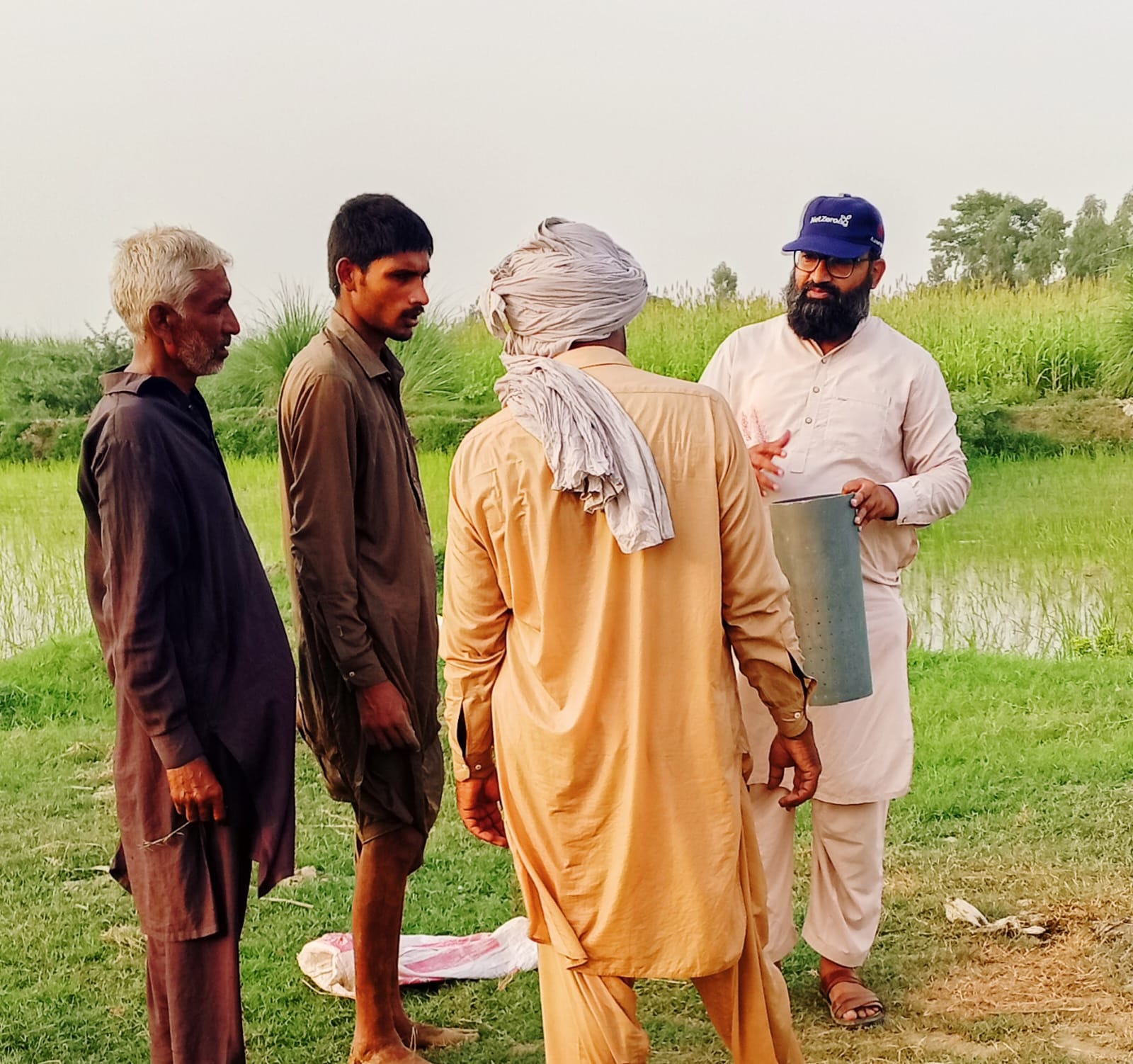 Four men standing and conversing outdoors in a green field, one holding a cylindrical metal object.