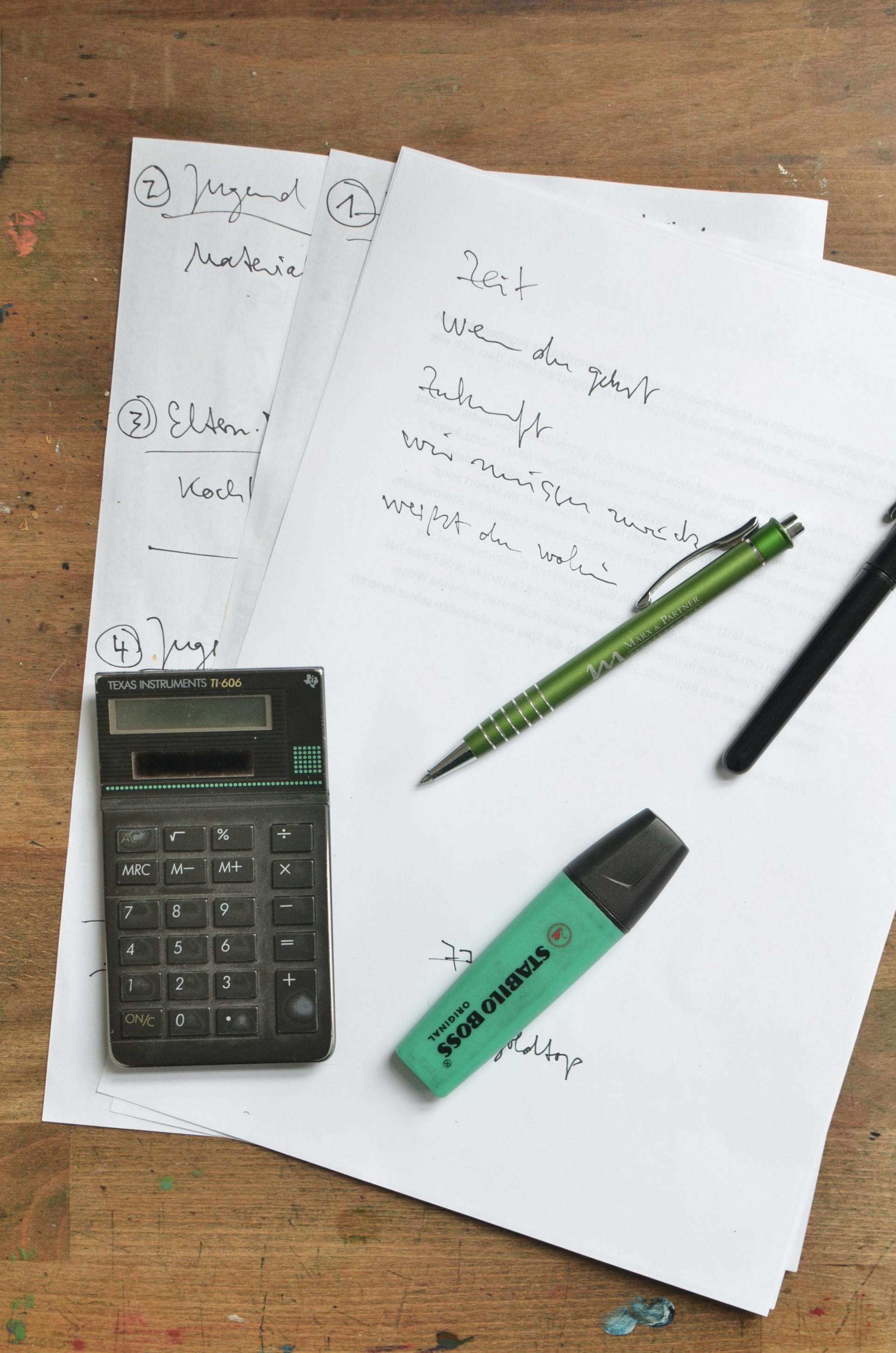 Schreibtisch mit Taschenrechner, Marker, Stiften und handbeschriebenen Blättern.