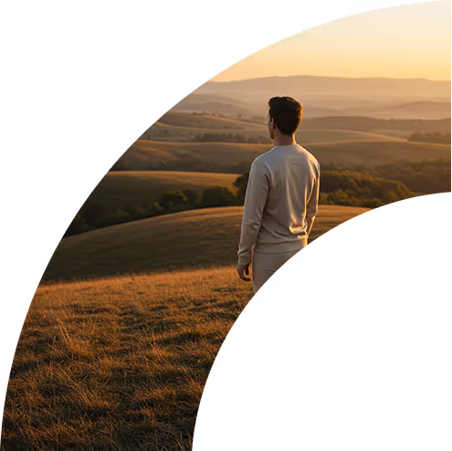 Man in light clothing standing on a grassy hill during sunset, overlooking rolling hills and trees.