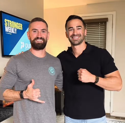 Two smiling men posing indoors, one wearing a gray shirt making a shaka hand sign and the other in a black shirt with a clenched fist.