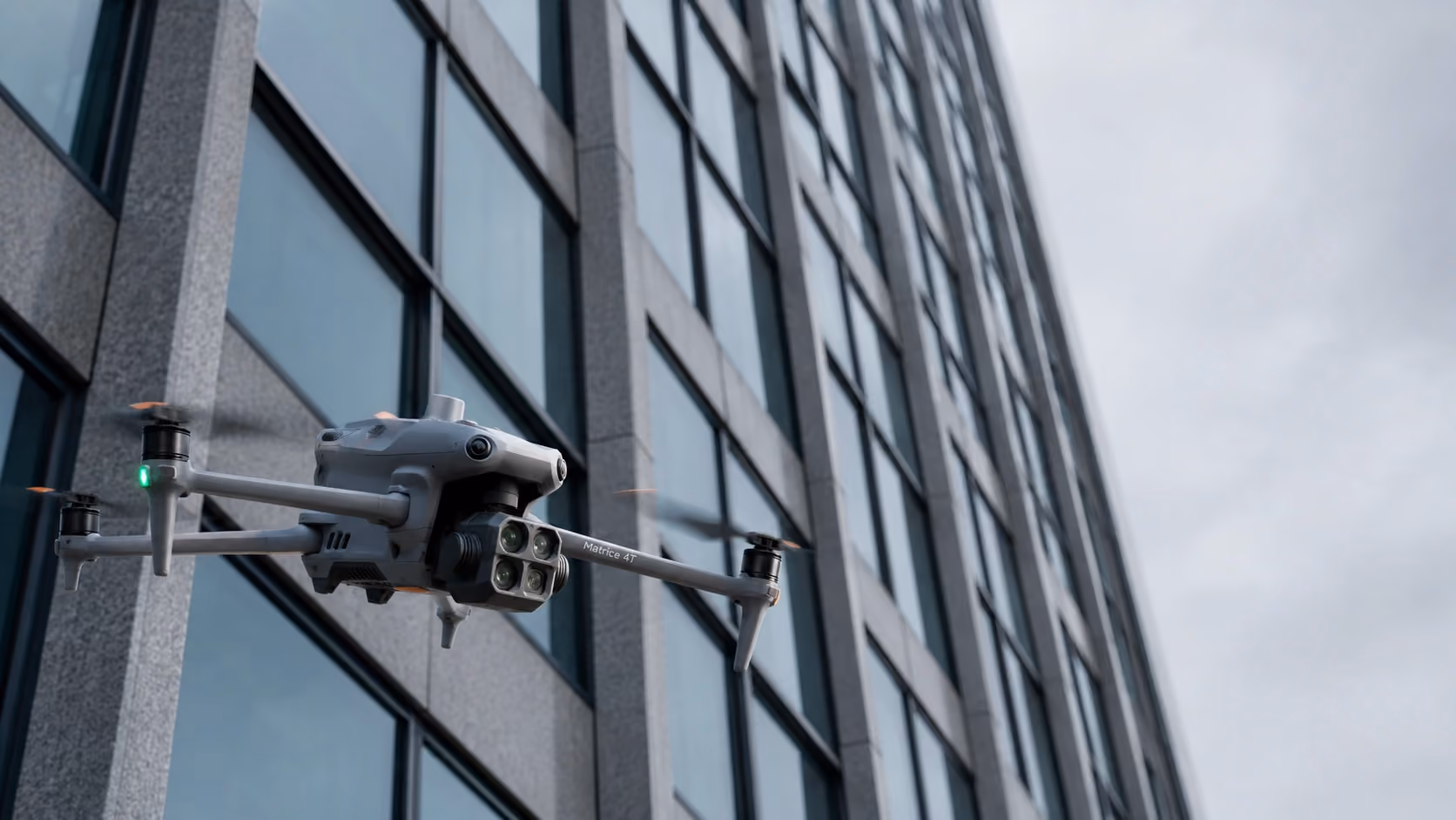 Graue Drohne mit vier Propellern fliegt vor einem modernen Bürogebäude mit Glasfront.