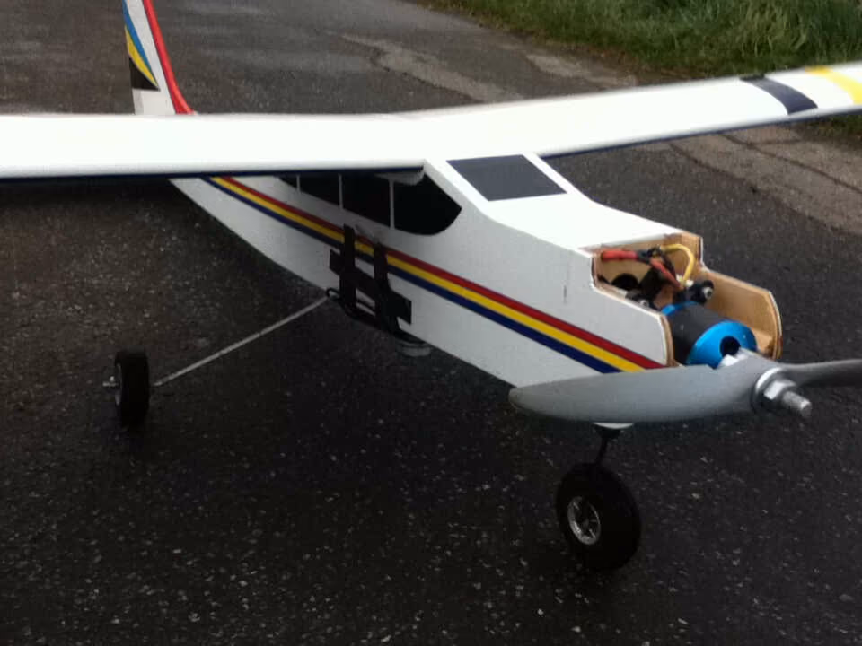 Weißes Modellflugzeug mit Verbrennungsmotor und Kamera unter dem Rumpf auf einem Weg.