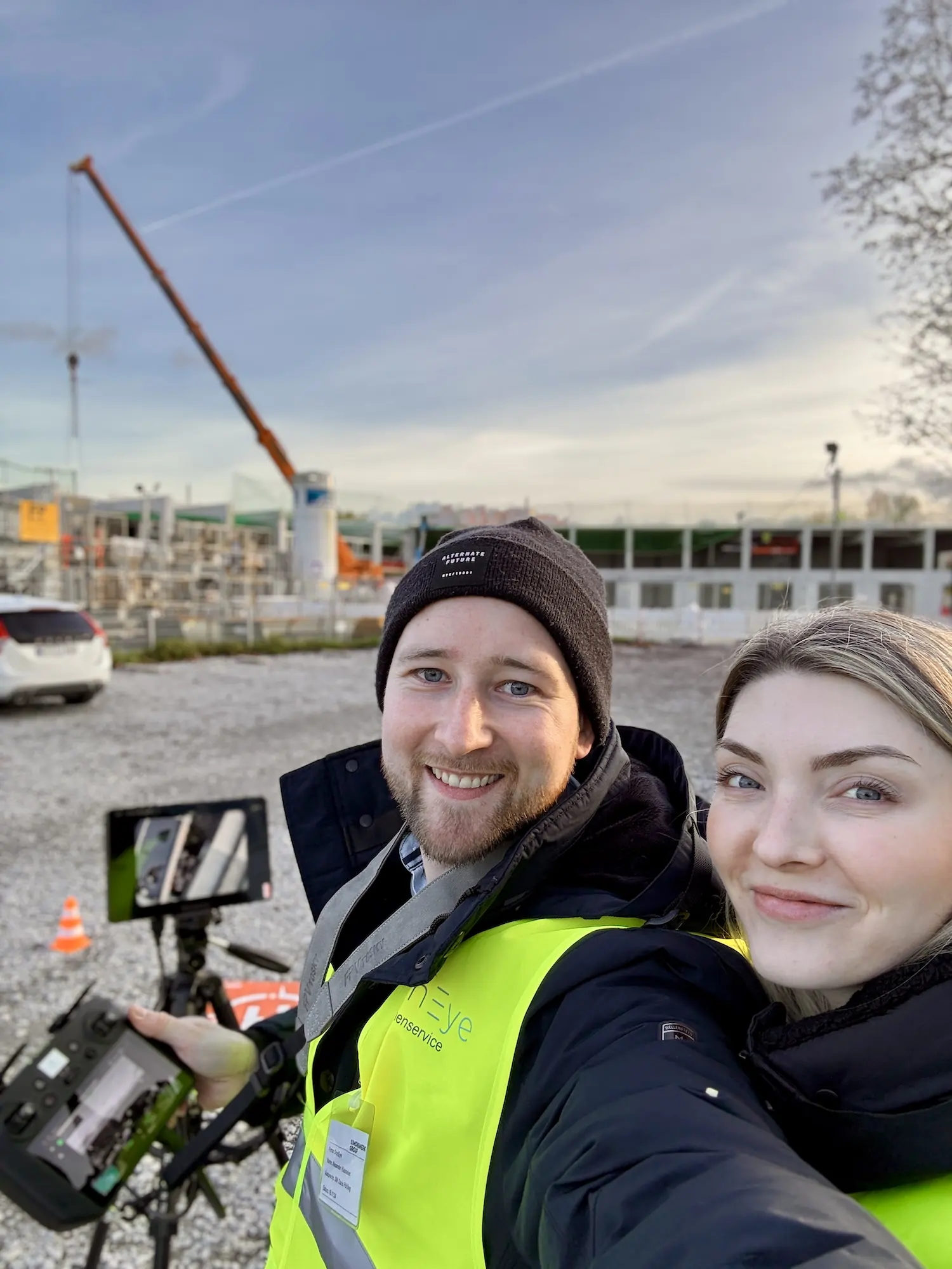 Alexander und Anna Klasmeier auf einer Baustelle, mit Drohnen-Fernsteuerung und Monitor, im Hintergrund ein Baukran