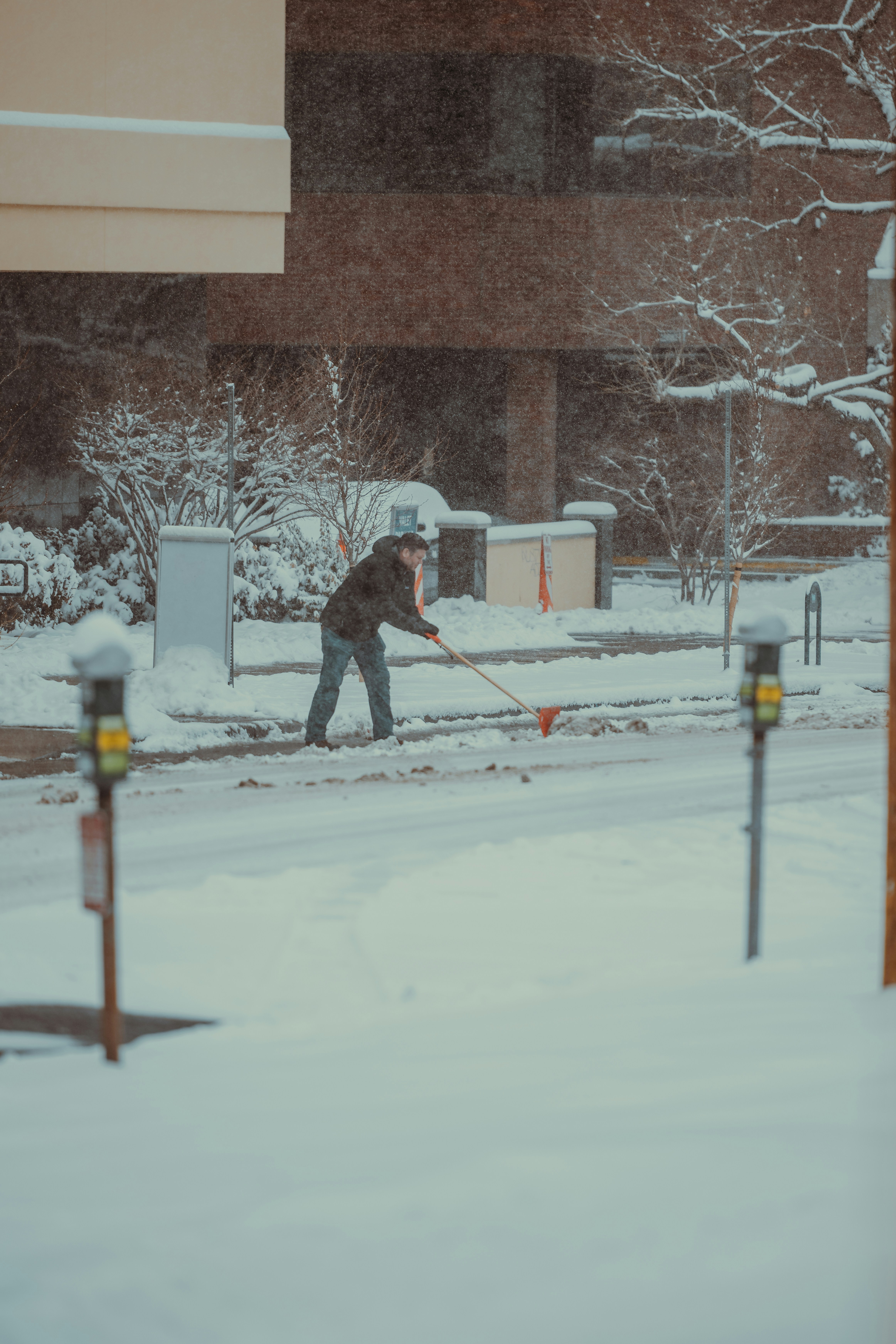 Mitarbeiter mit Warnweste schaufelt Schnee vor einem Gebäude mit einer orangen Schneeschippe