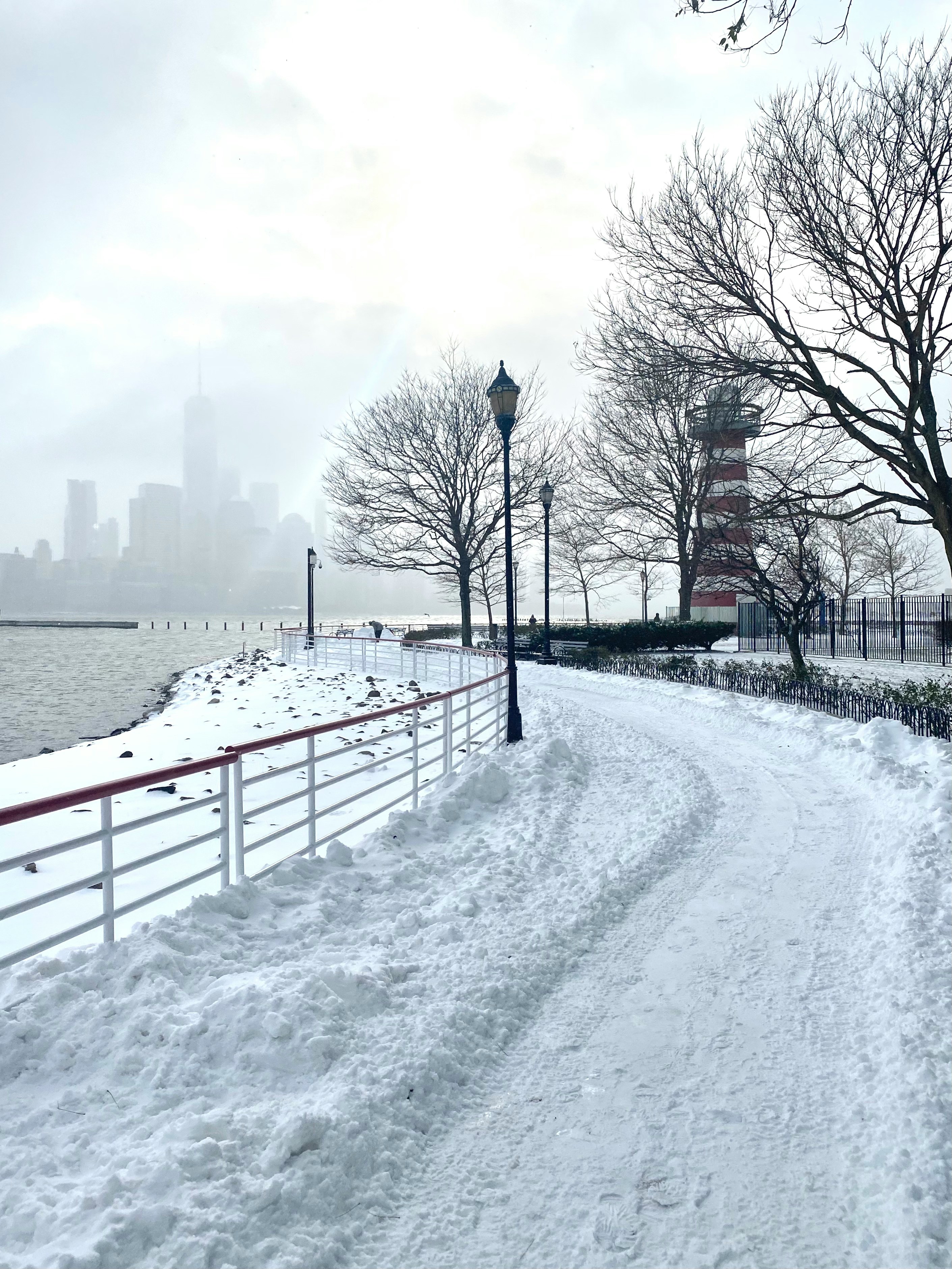 Verschneiter Uferweg mit Leuchtturm und neblige New Yorker Skyline im Winterschnee