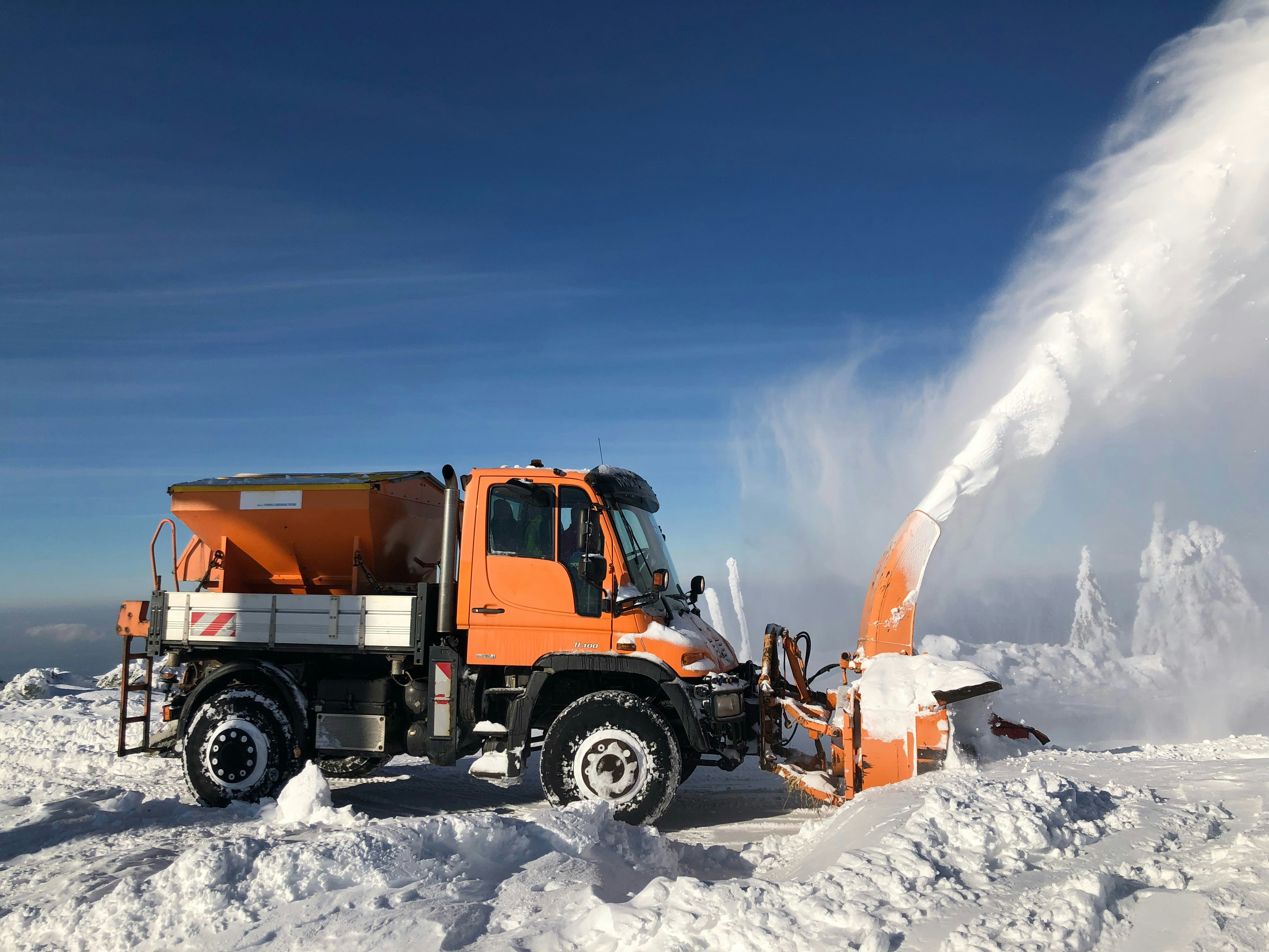 Orangefarbener Unimog mit Schneefräse schleudert Schnee in winterlicher Berglandschaft unter blauem Himmel