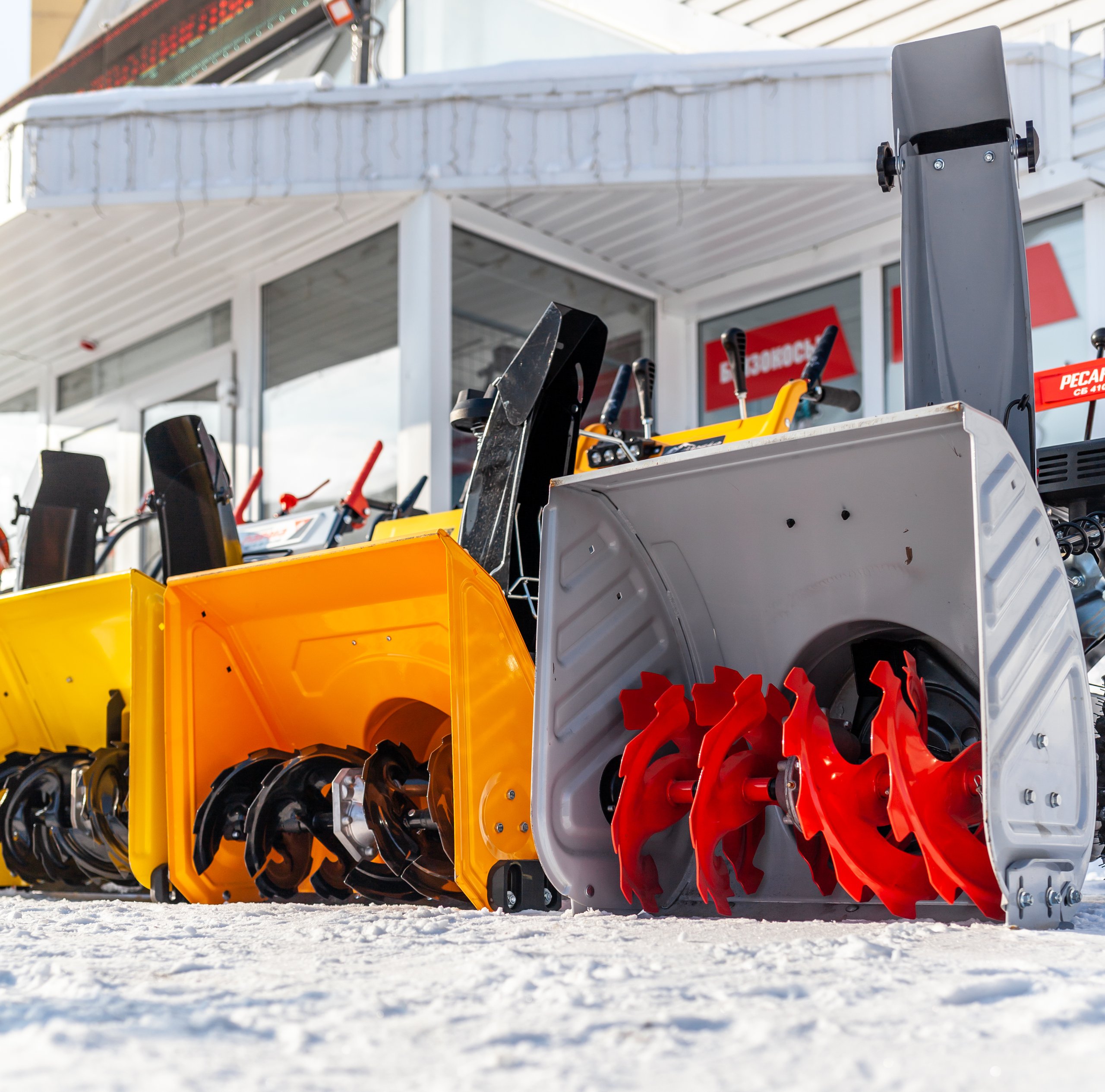 Mehrere gelbe, orange und graue Schneefräsen stehen in einer Reihe vor einem Geschäft im Schnee