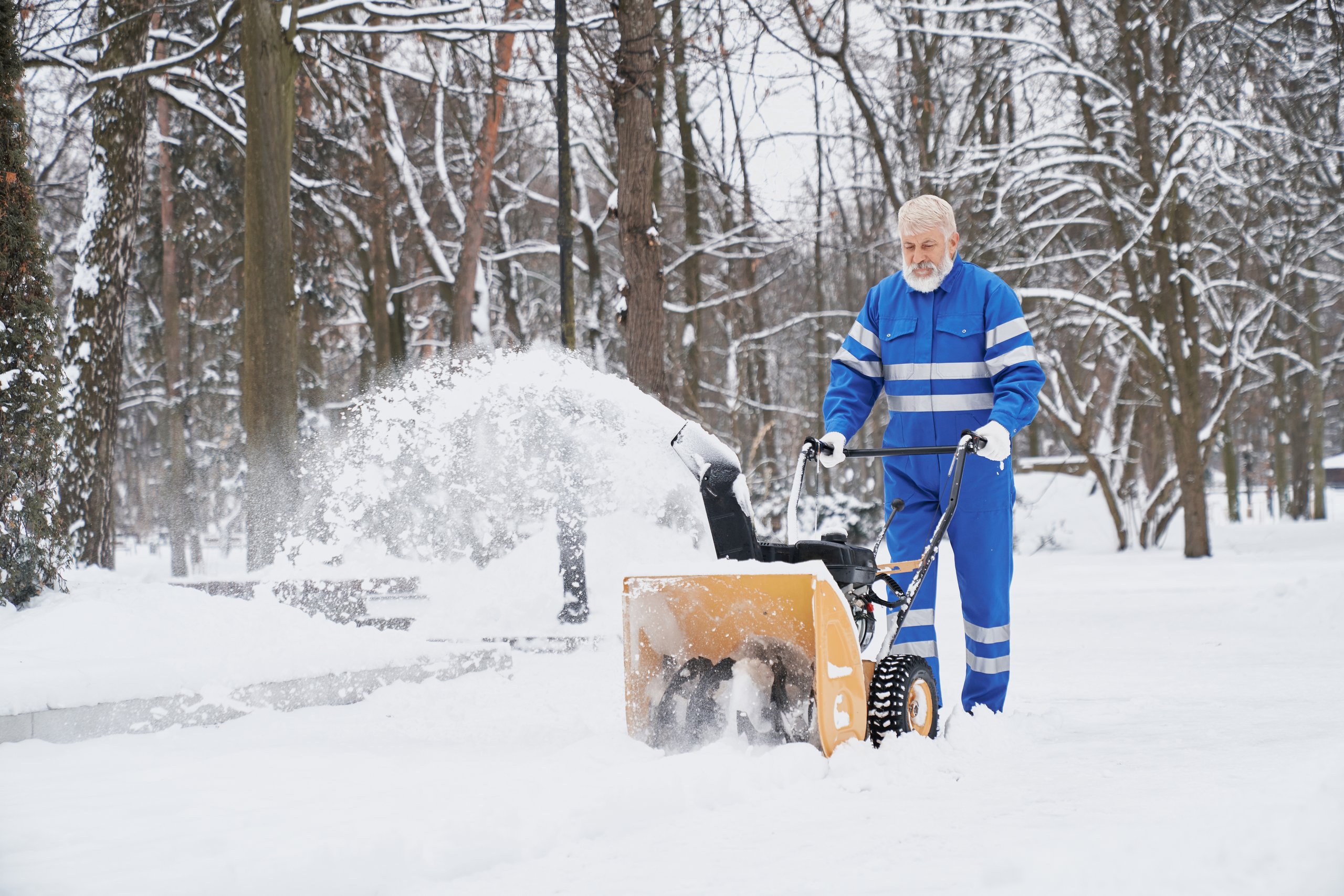 Mann in blauer Arbeitskleidung räumt mit einer Schneefräse einen verschneiten Weg im Park