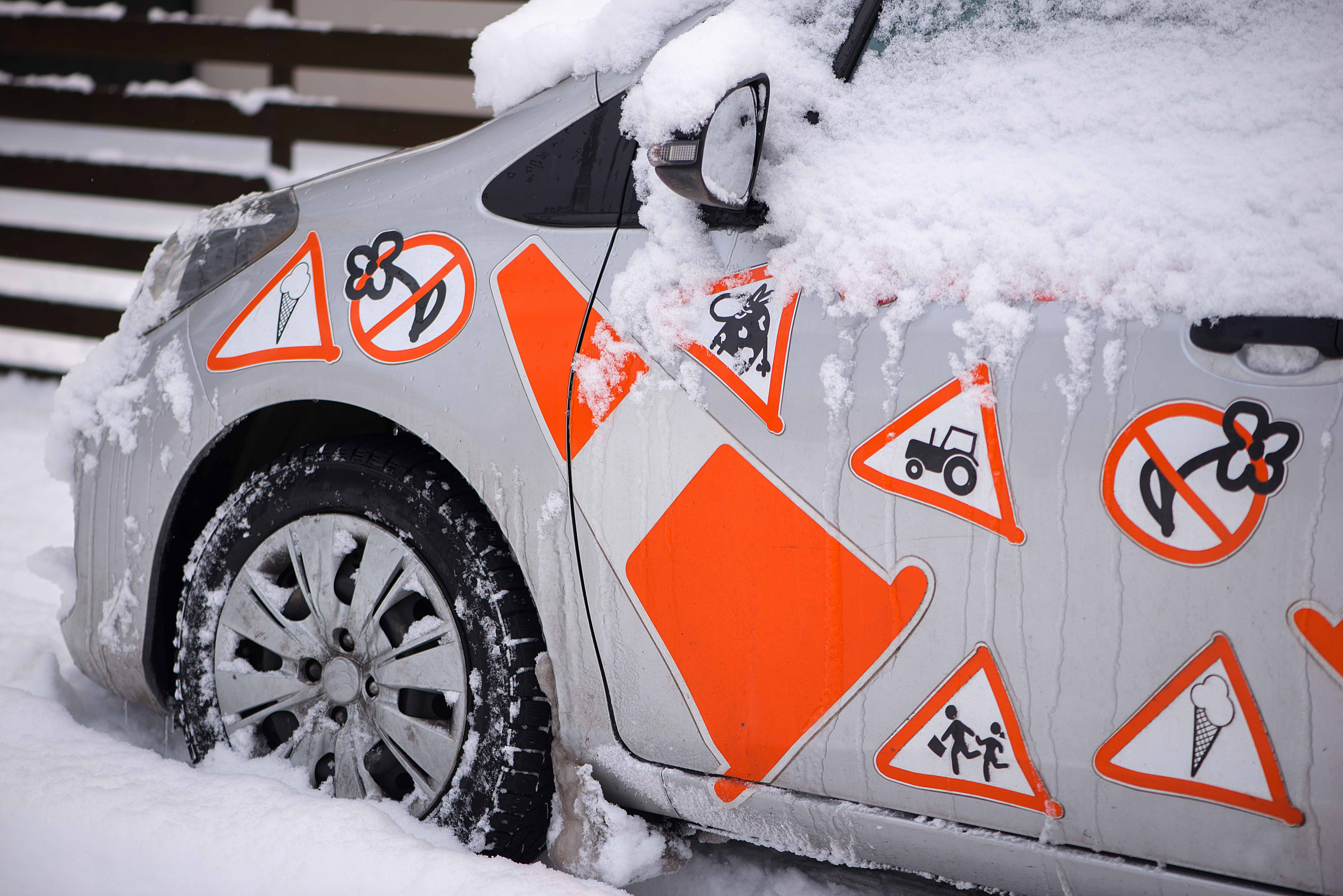 Schneebedecktes Auto mit humorvollen orangefarbenen Warnschildern für Eis, Fahrrad, Kuh, Traktor und spielende Kinder auf der Fahrertür