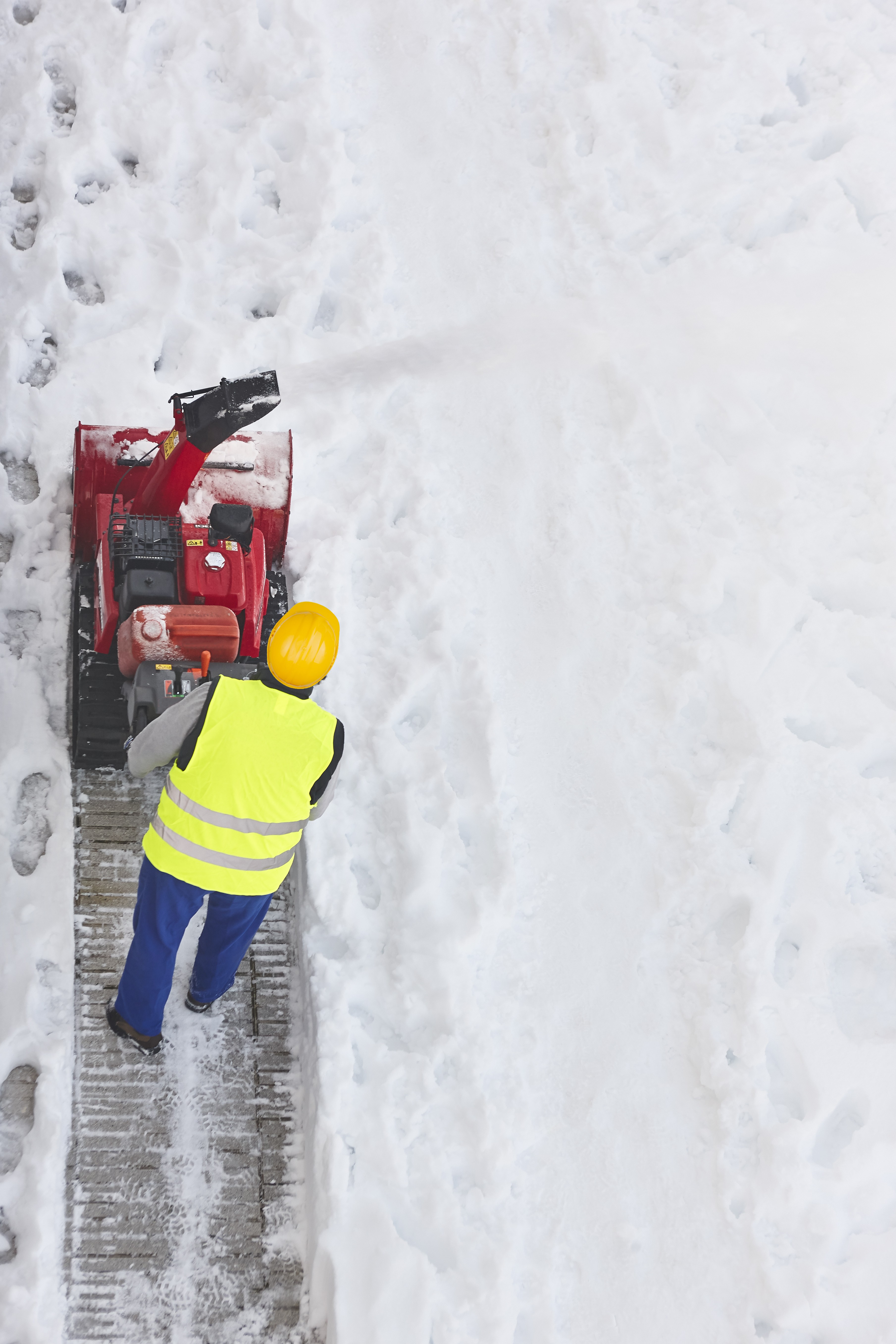 Bauarbeiter mit gelbem Helm und Warnweste bedient eine Schneefräse auf einem verschneiten Gehweg von oben fotografiert
