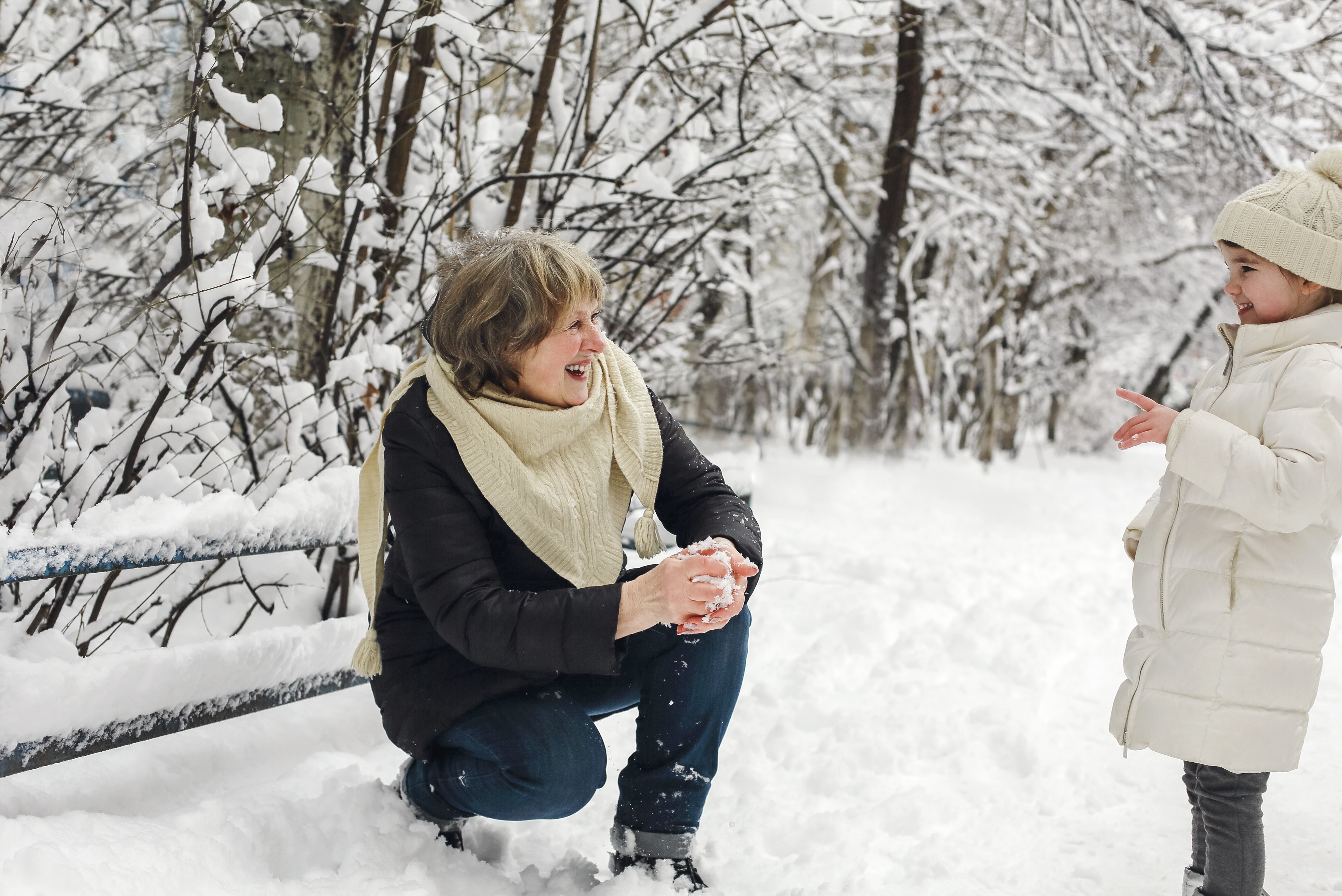 Lachende Großmutter formt kniend einen Schneeball und schaut ein kleines Mädchen im weißen Wintermantel im verschneiten Park an