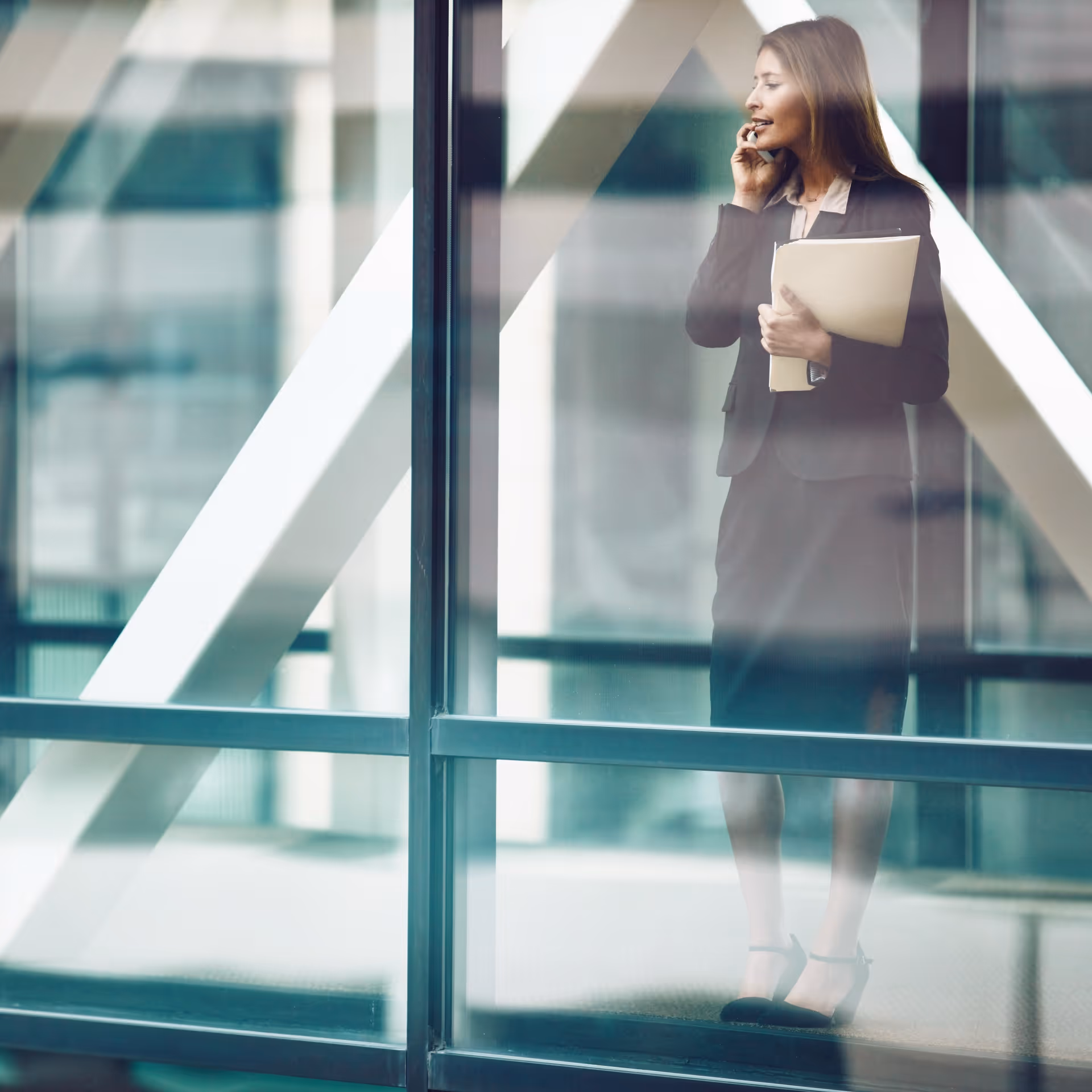 Businesswoman in a suit talking on phone and holding documents inside an office with glass windows.