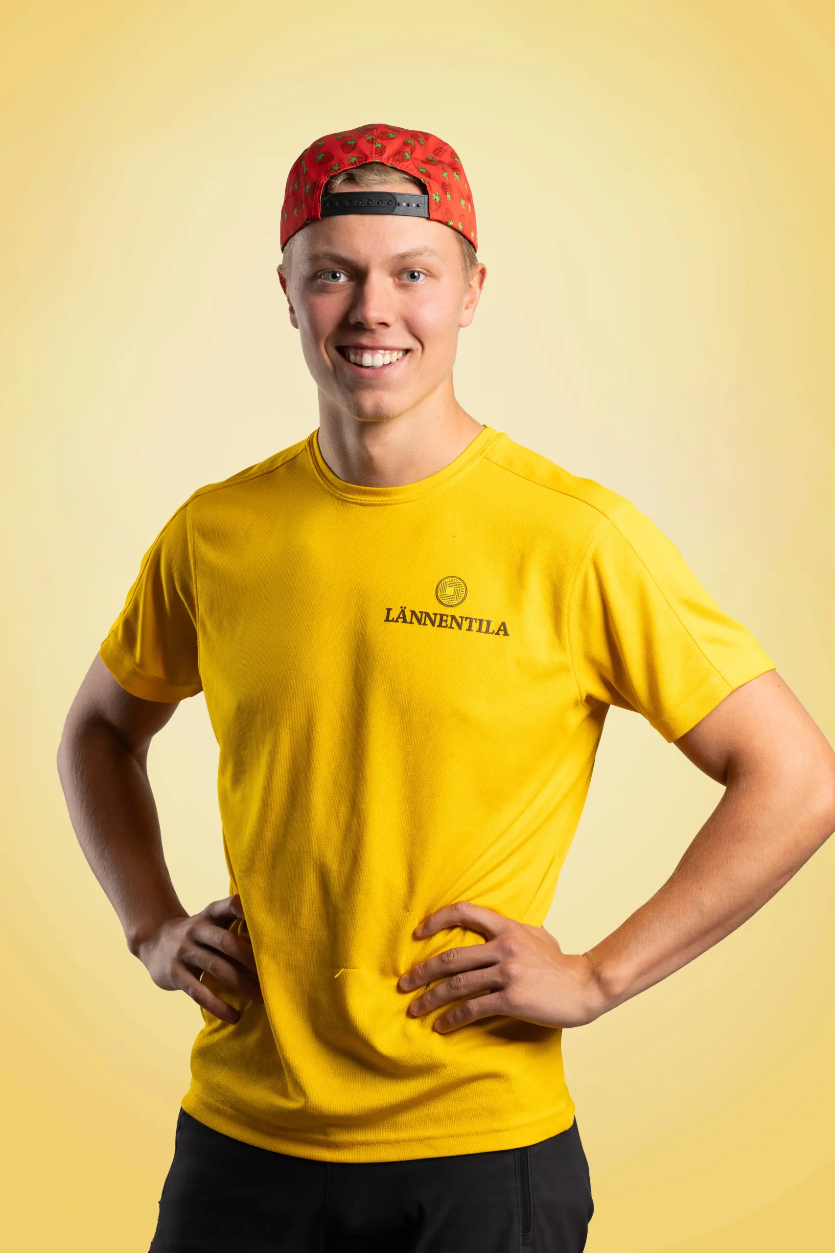 Nuori mies hymyilee kameralle, kädet lanteilla. Hänellä on päässään takaperin punainen mansikkakuvioinen lippalakki ja yllään keltainen t-paita, jossa on musta LÄNNENTILA-logo. Tausta on vaaleankeltainen.