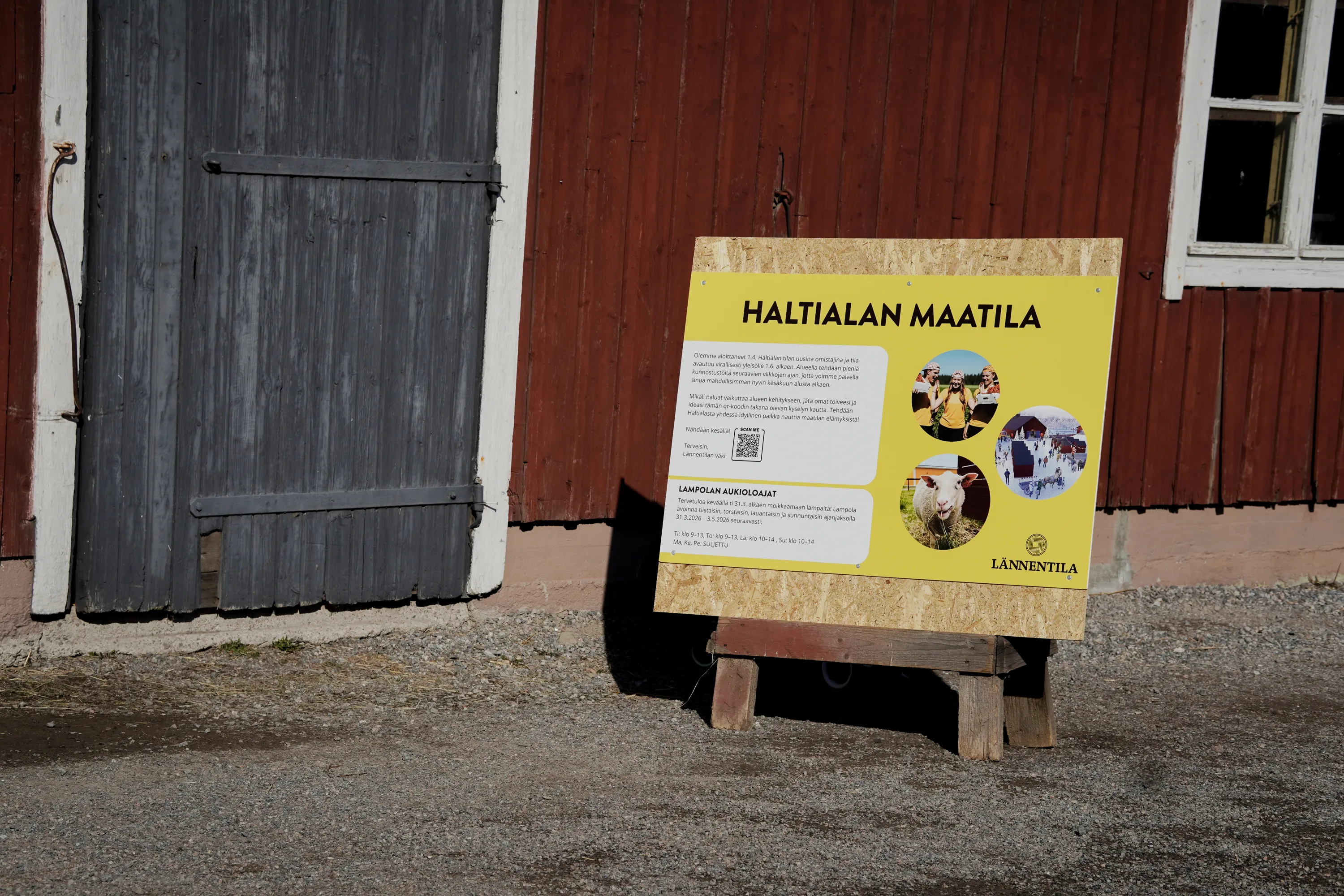Keltainen kyltti puujalustalla seisoo punaisen puurakennuksen edessä. Rakennuksessa on tummanharmaa puuovi vasemmalla ja valkoinen ikkunanpuitteet oikealla. Kyltissä on musta teksti "HALTIALAN MAATILA", suomenkielistä tietoa, QR-koodi ja kolme pyöreää kuvaa: hymyileviä ihmisiä, lammas syömässä heinää ja talvinen kylämaisema. Maa on soraa. Kohtaus on kirkkaasti auringon valaisema.