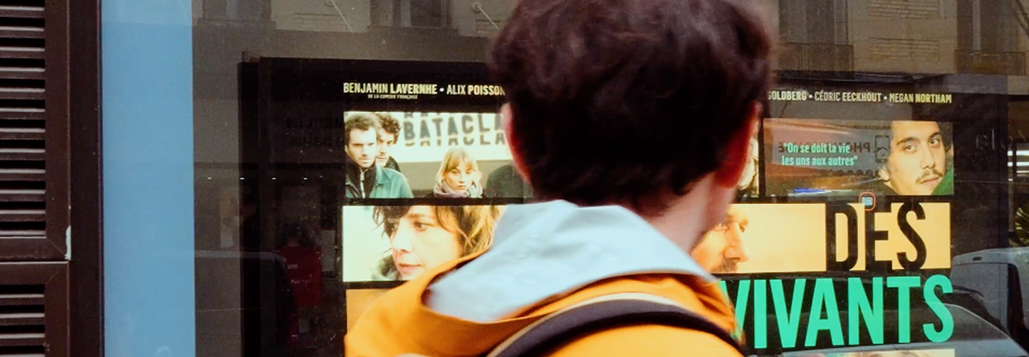 Un jeune homme vu de dos portant une veste orange devant une affiche de film affichée dans une vitrine.