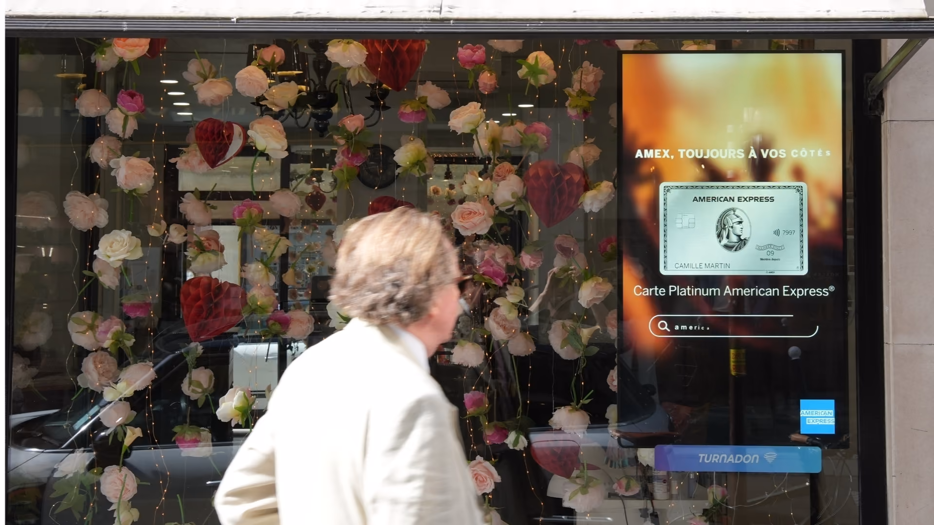 Une vitrine fleurie avec une publicité pour la carte Platinum American Express à côté d'un homme en manteau clair.
