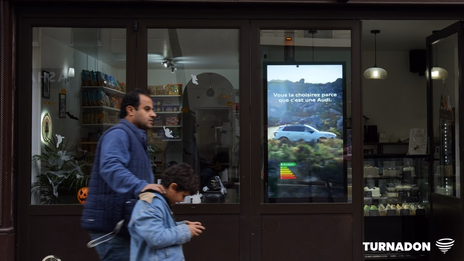 Un homme et un enfant passent devant une vitrine où s’affiche une publicité Audi en français.
