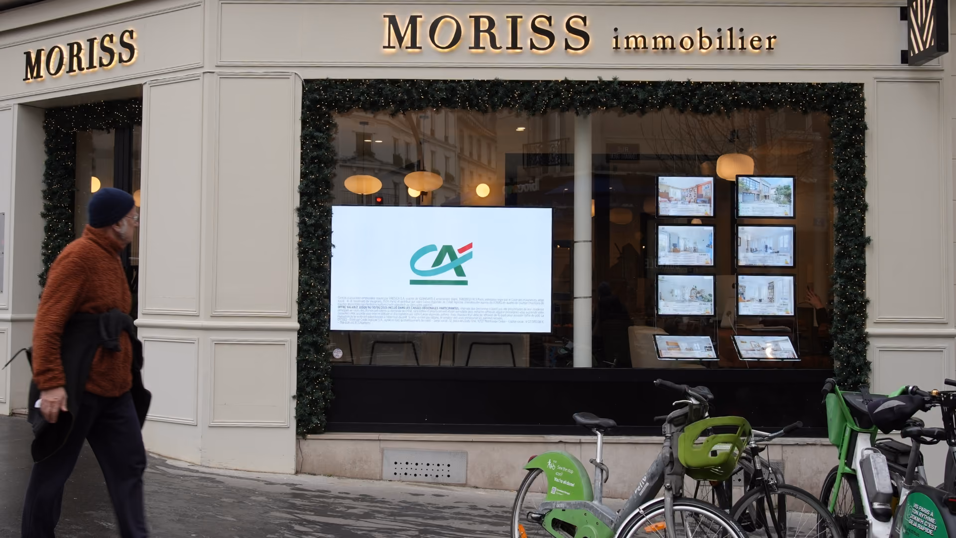 Vitrine d'agence immobilière MORISS avec écrans d'annonces et vélos en stationnement devant.