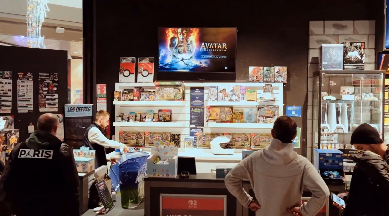 Avec quatre clients, un employé sert au comptoir d'un magasin de jeux vidéo avec une pub TV pour Avatar.