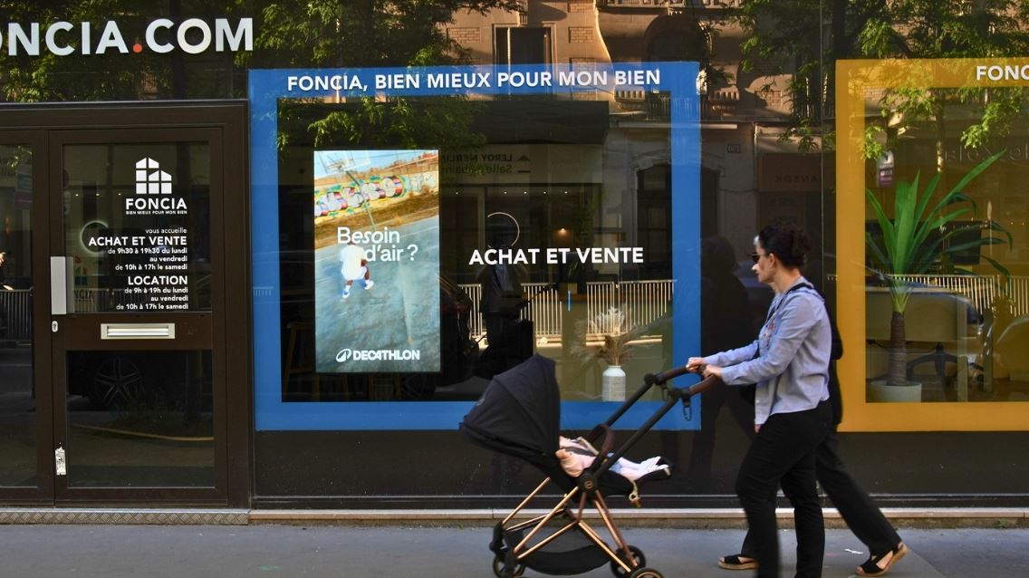 Deux femmes à poussette passent devant une vitrine et regardent l'écran digital avec une publicité pour Décathlon