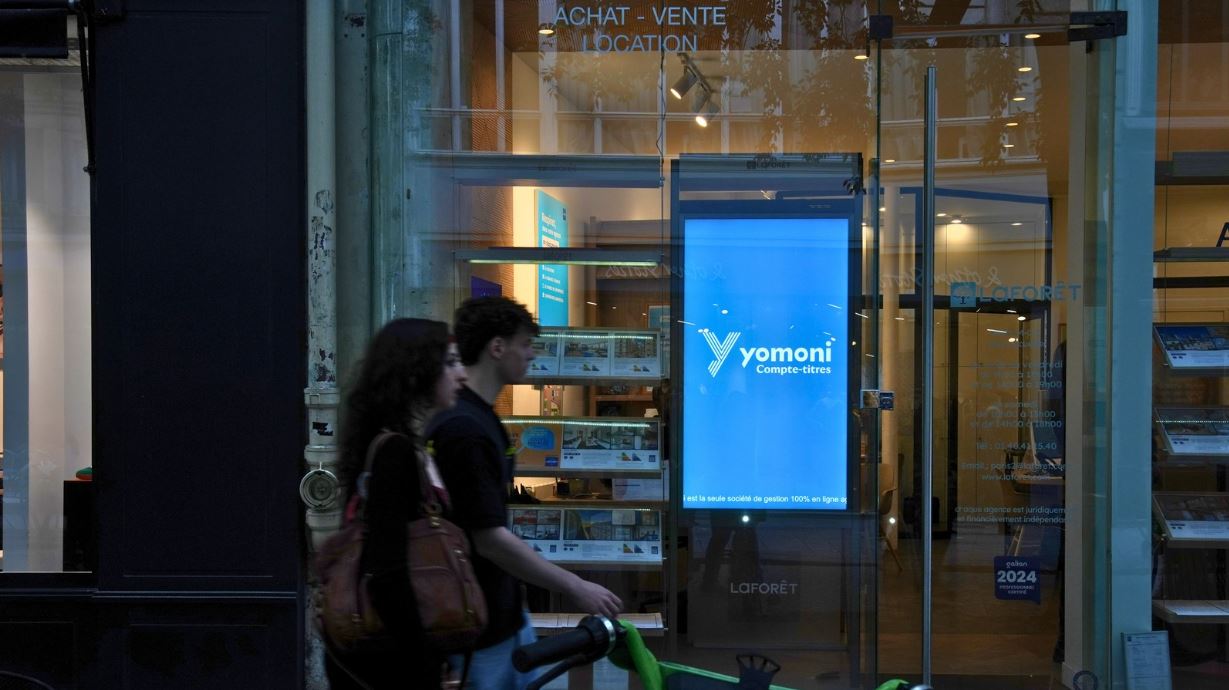 Un jeune couple passe devant une agence immobilière et regarde la campagne publicitaire de Yomoni