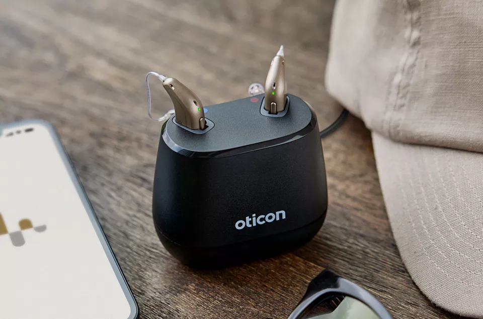 Pair of Oticon hearing aids resting in a black charging dock on a wooden surface alongside a smartphone and part of a fabric cap.