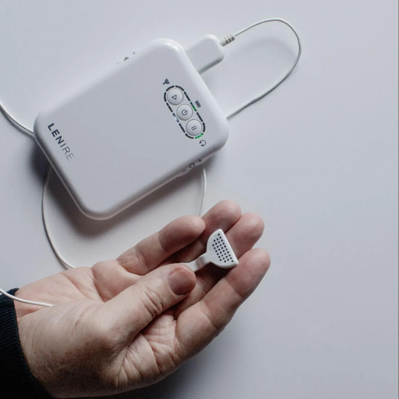 Hand holding a small white device with perforated surface connected to a white rectangular electronic unit labeled LENIRE.
