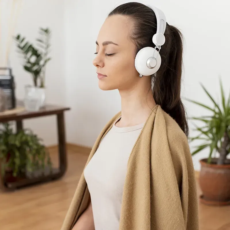 Woman with eyes closed wearing white headphones and a beige shawl, sitting indoors with plants in the background.