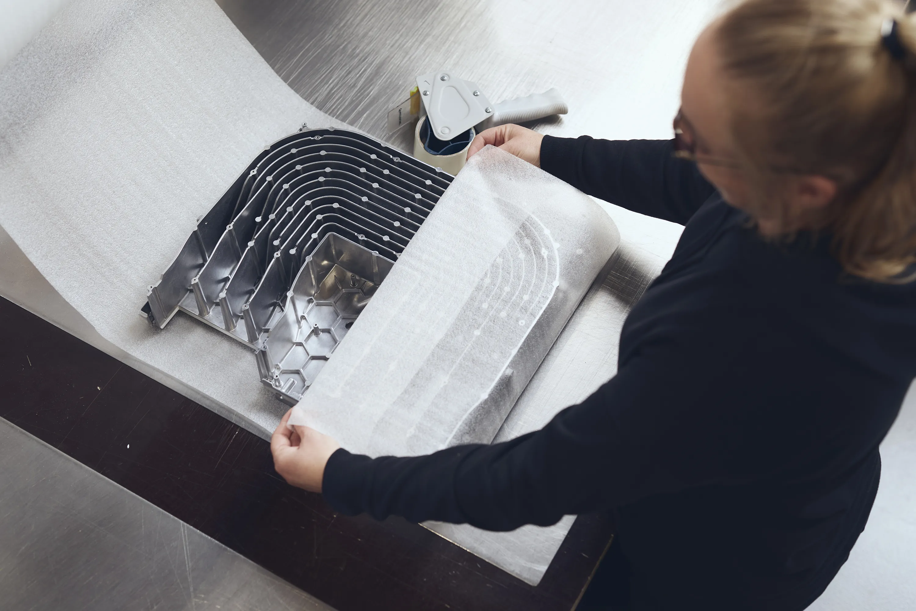Person wrapping stack of curved metal parts with protective foam sheet on metal table.