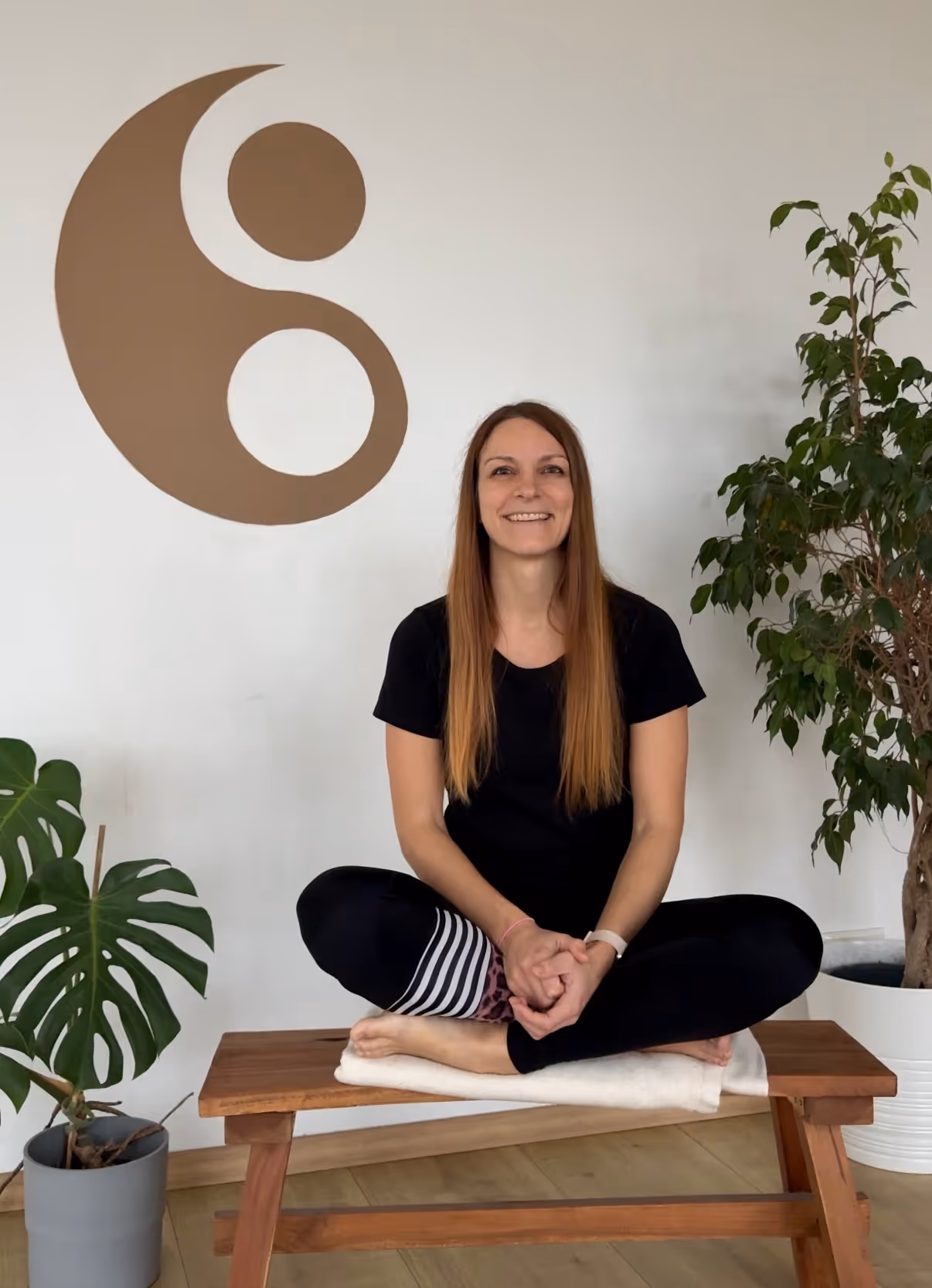 Jule, Yogalehrerin bei Nalu Yoga Rostock, sitzt im Schneidersitz lächelnd auf einer Bank im Studio vor dem Yin-Yang-Symbol
