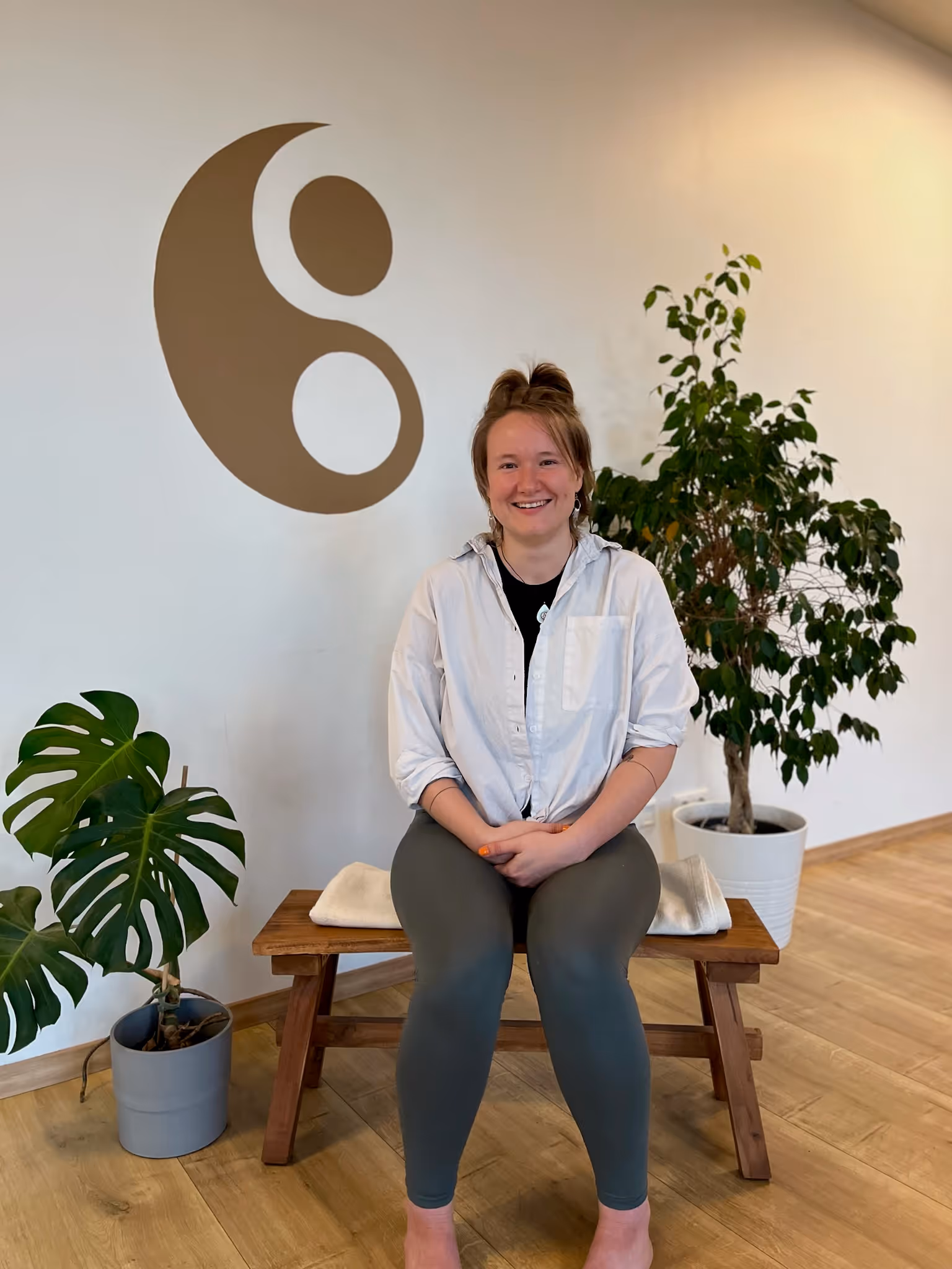 Leni, Yogalehrerin bei Nalu Yoga Rostock, sitzt lächelnd auf einer Bank im Studio vor dem Yin-Yang-Symbol