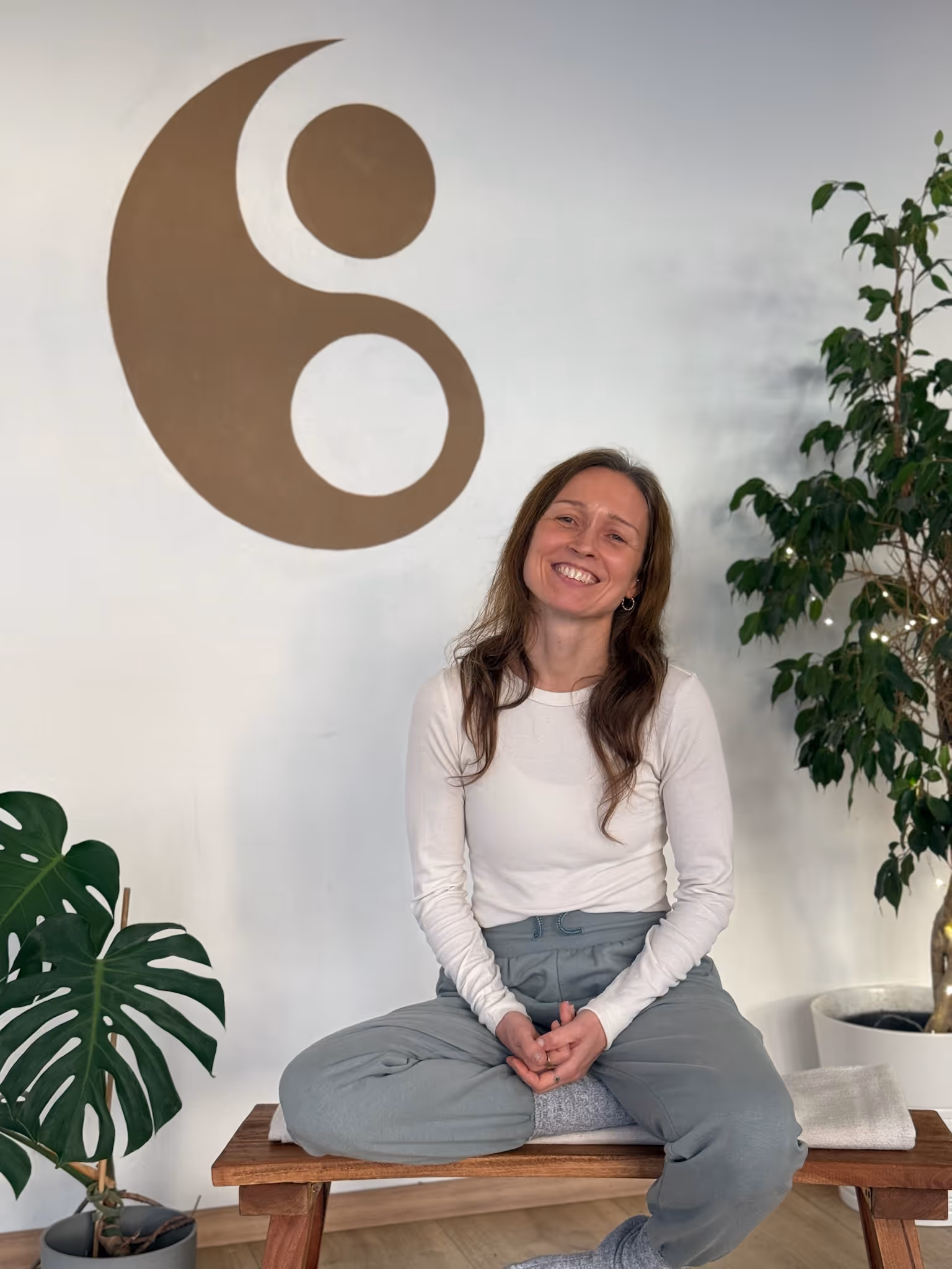 Anika, Yogalehrerin bei Nalu Yoga Rostock, sitzt im Schneidersitz lächelnd auf einer Bank im Studio vor dem Yin-Yang-Symbol