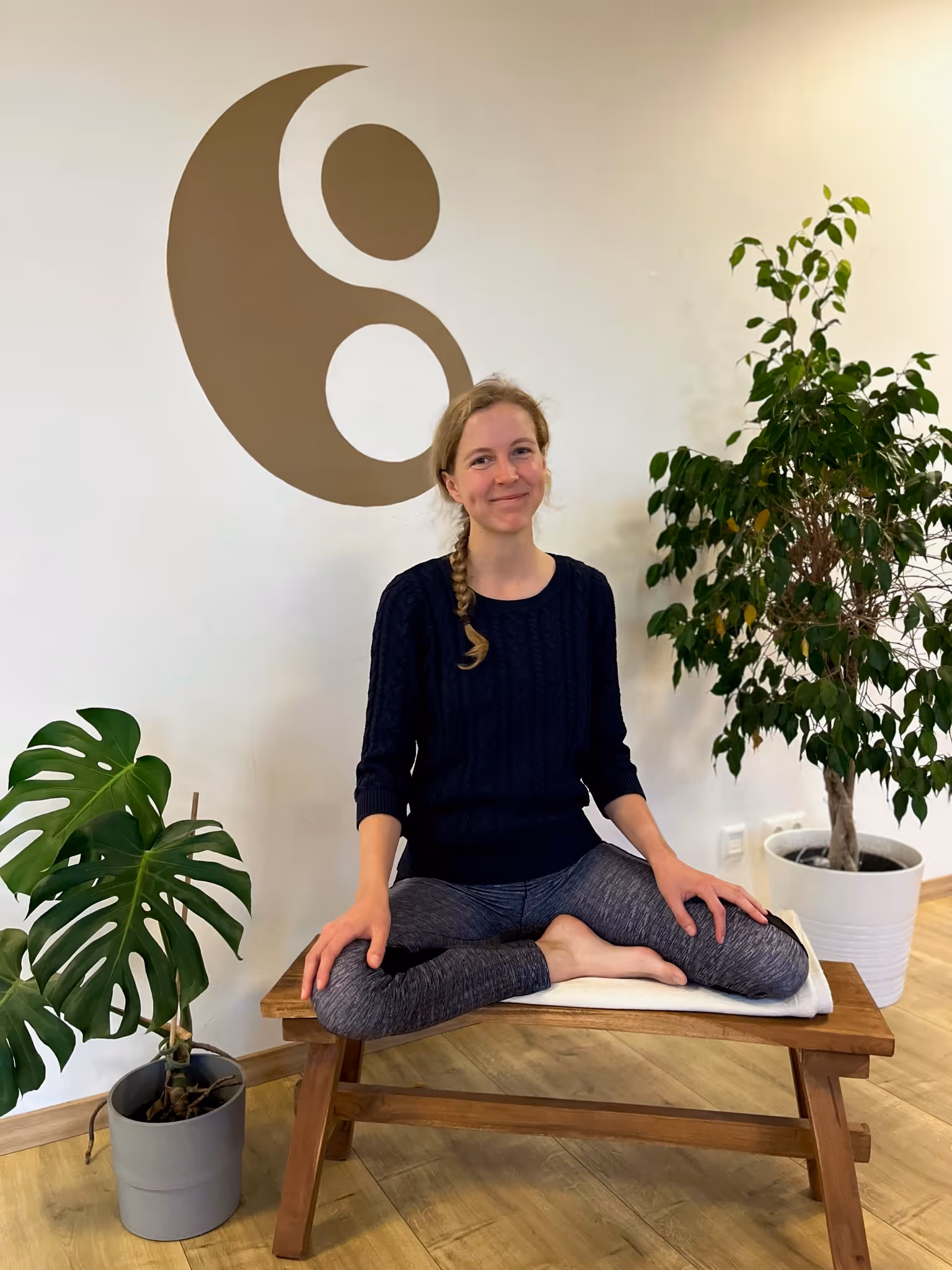 Jenny, Yogalehrerin bei Nalu Yoga Rostock, sitzt im Schneidersitz lächelnd auf einer Bank im Studio vor dem Yin-Yang-Symbol