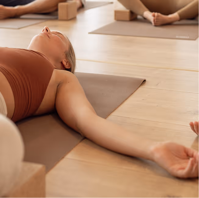 Frau in Yin Yoga Entspannungspose auf Matte bei Nalu Yoga Rostock, umgeben von Yogablöcken und weiteren Kursteilnehmern