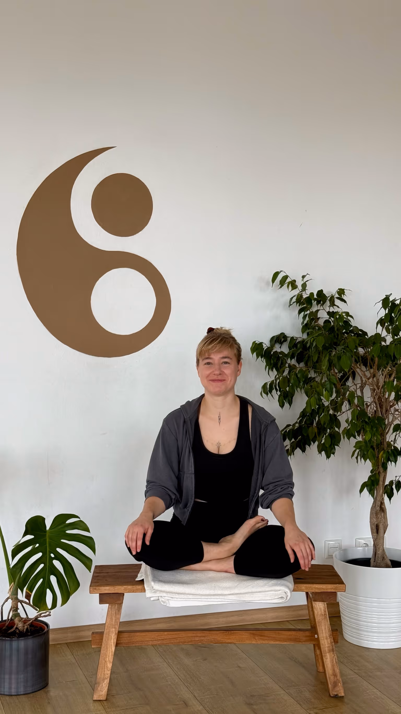 Johanna, Yogalehrerin bei Nalu Yoga Rostock, sitzt im Schneidersitz lächelnd auf einer Bank im Studio vor dem Yin-Yang-Symbol