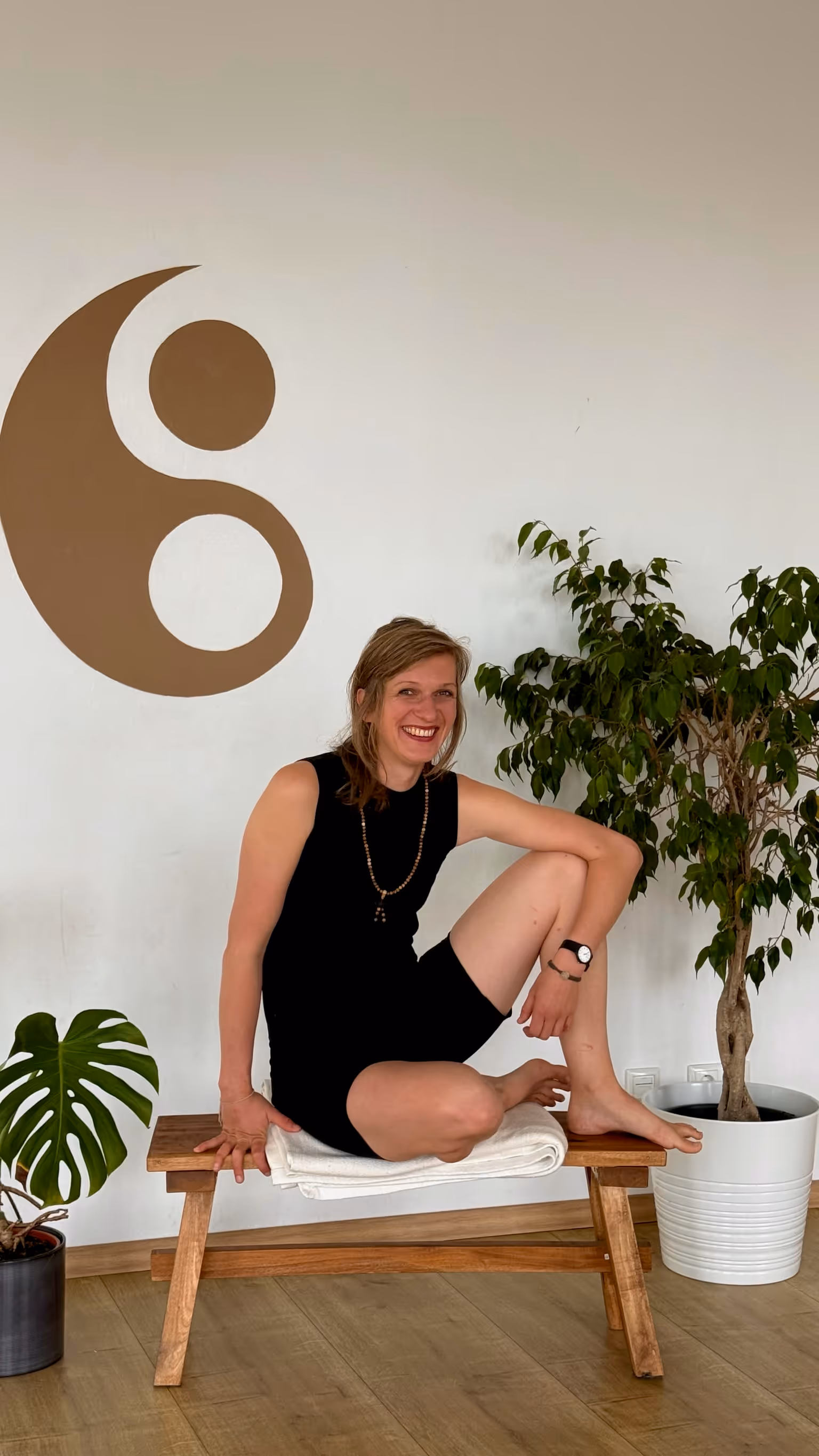 Juliane Yogalehrerin bei Nalu Yoga Rostock, sitzt im Schneidersitz lächelnd auf einer Bank im Studio vor dem Yin-Yang-Symbol