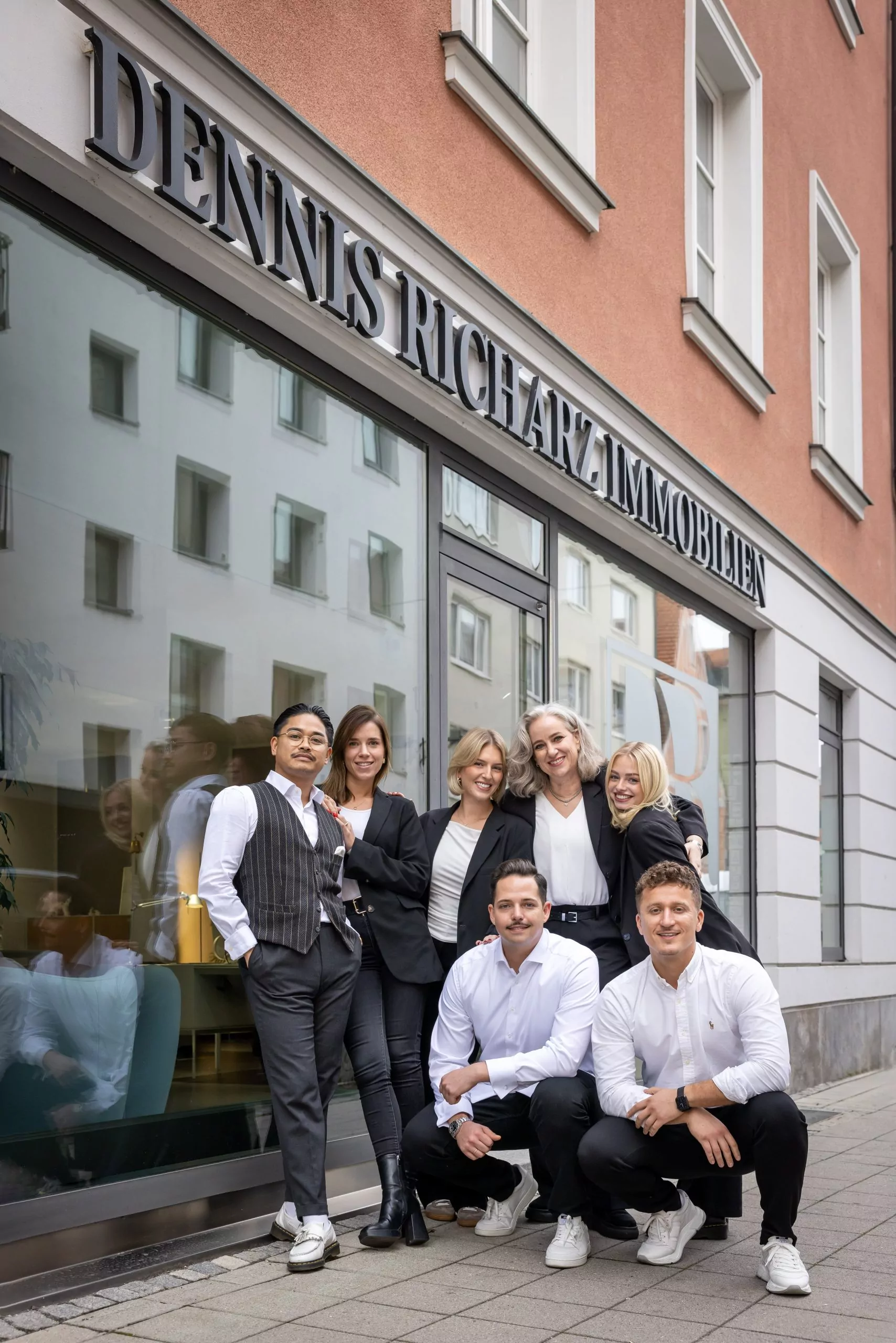 Sechs Personen stehen und hocken vor einem Büro mit Schild Dennis Richarz Immobilien.