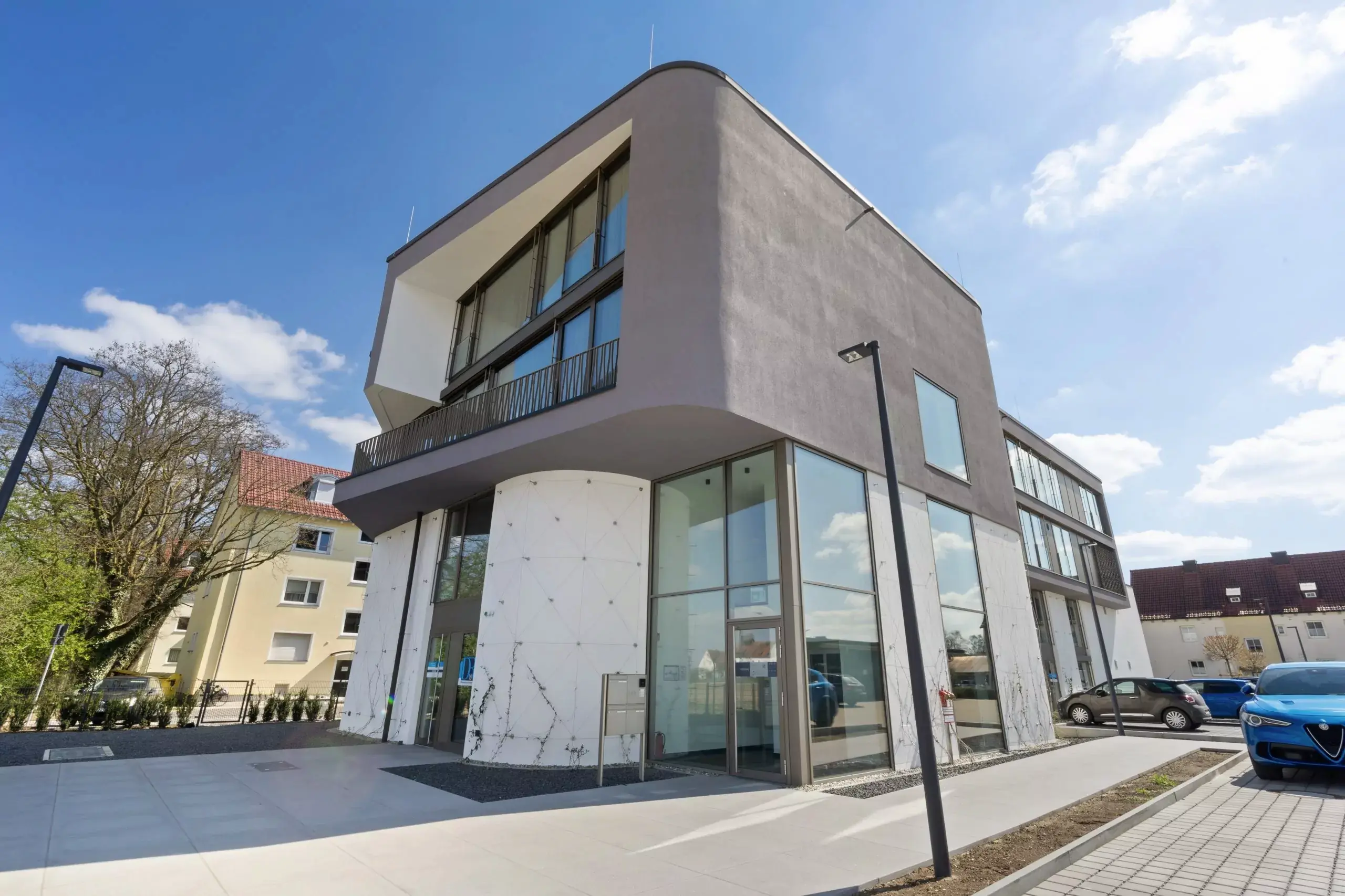 Modernes Bürogebäude mit großen Glasfenstern und geparkten Autos davor unter blauem Himmel.
