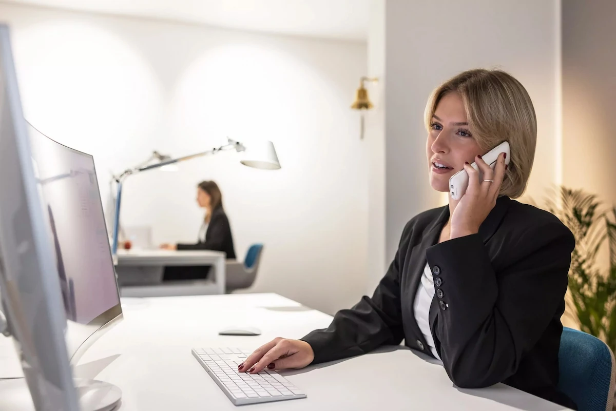 Frau im Büro am Computer spricht am Telefon, weitere Person im Hintergrund am Schreibtisch.