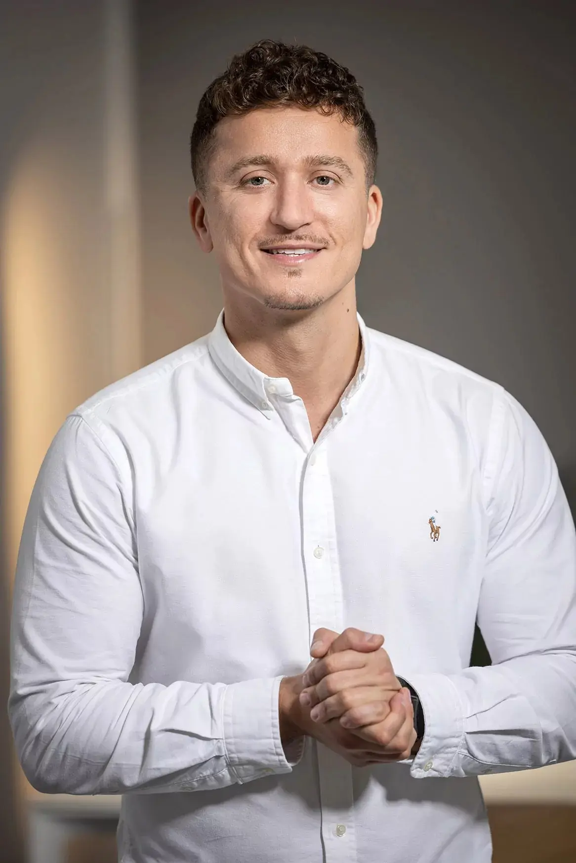 Ein Mann mit weißem Hemd steht lächelnd mit verschränkten Händen vor neutralem Hintergrund.