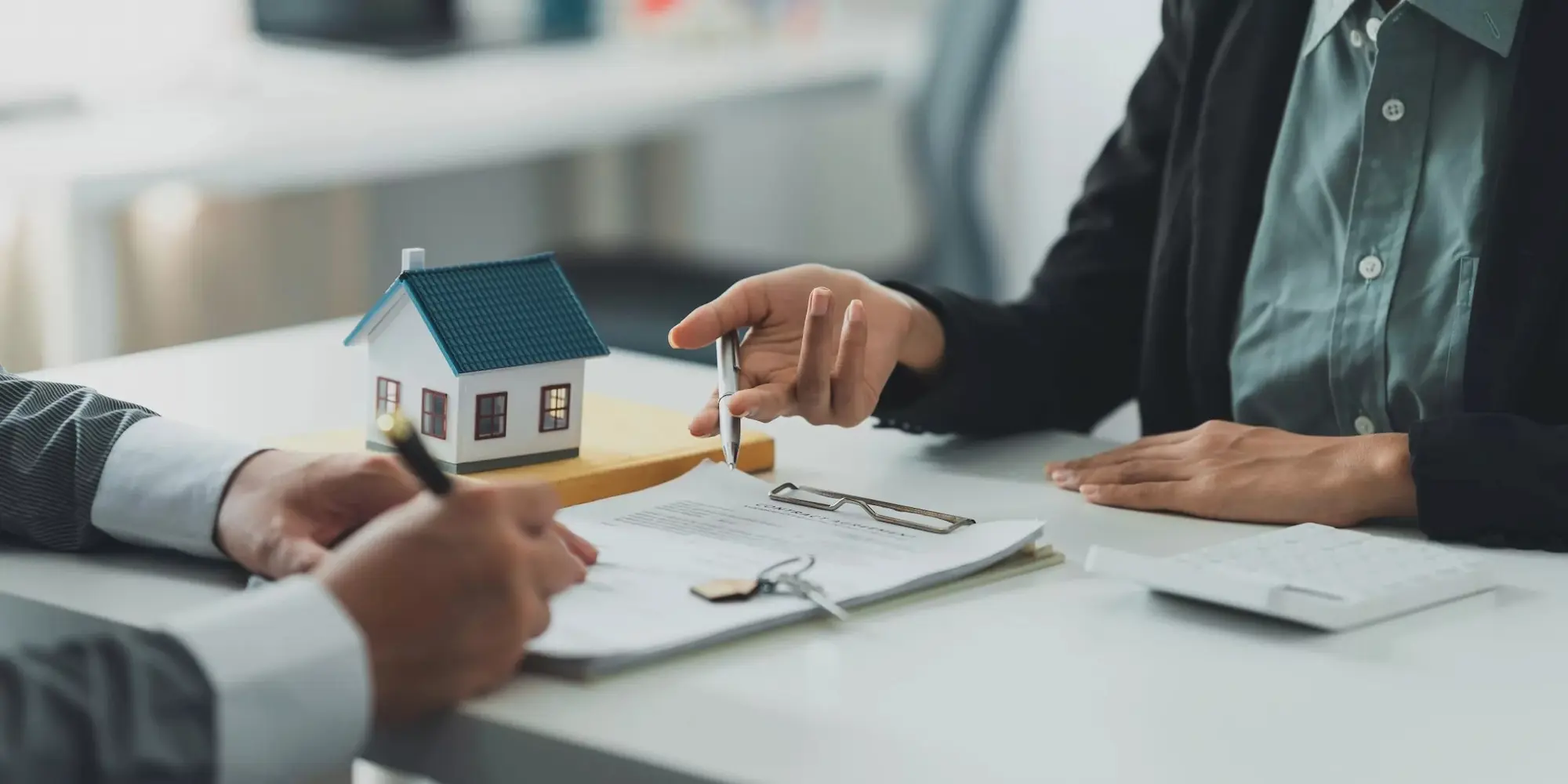 Zwei Personen sitzen am Tisch, eine hält einen Stift, daneben ein Modellhaus und Dokumente.