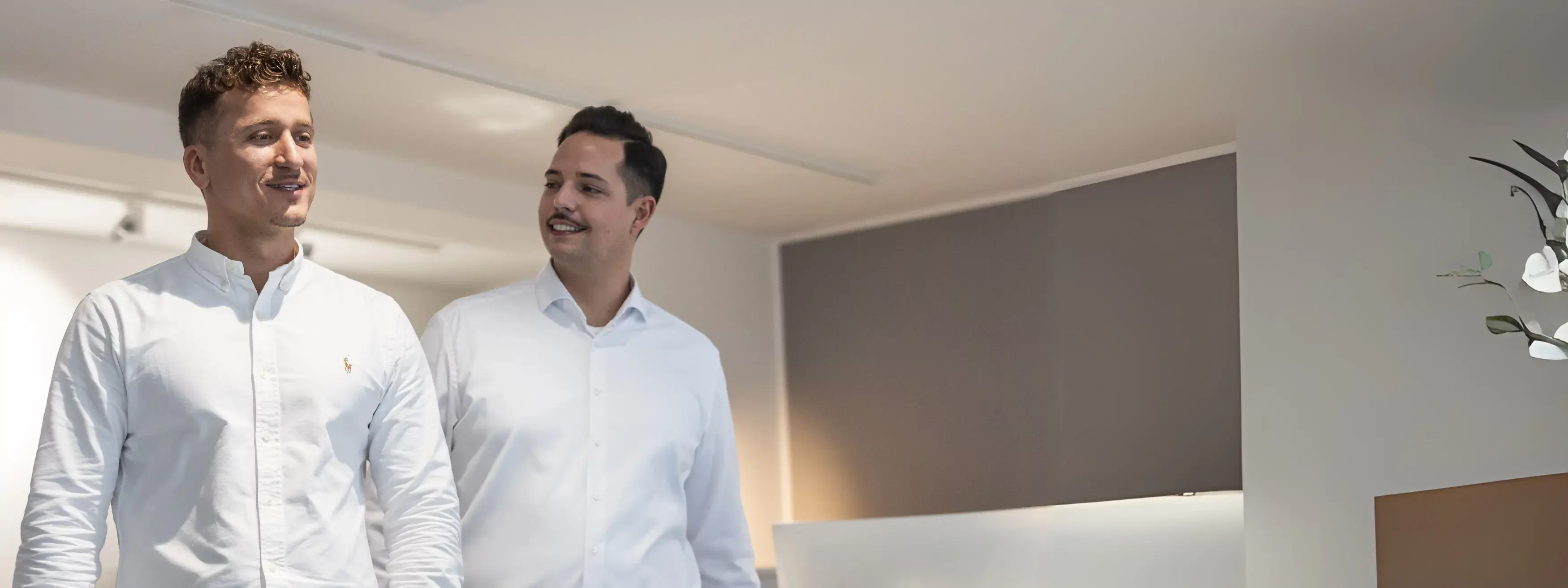 Zwei Männer in weißen Hemden unterhalten sich und lächeln in einem hellen Büro.