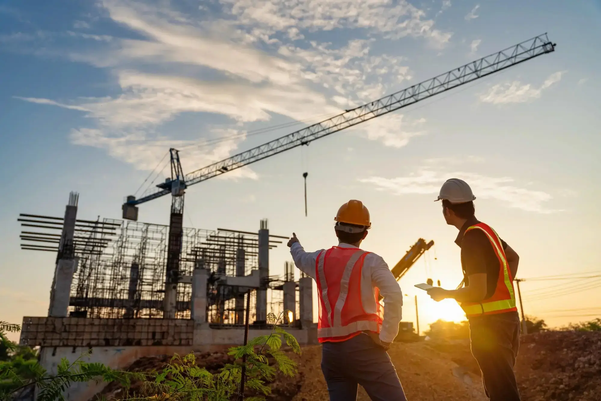 Zwei Bauarbeiter mit Schutzhelmen und Westen besprechen Baustelle mit Kran im Sonnenuntergang.