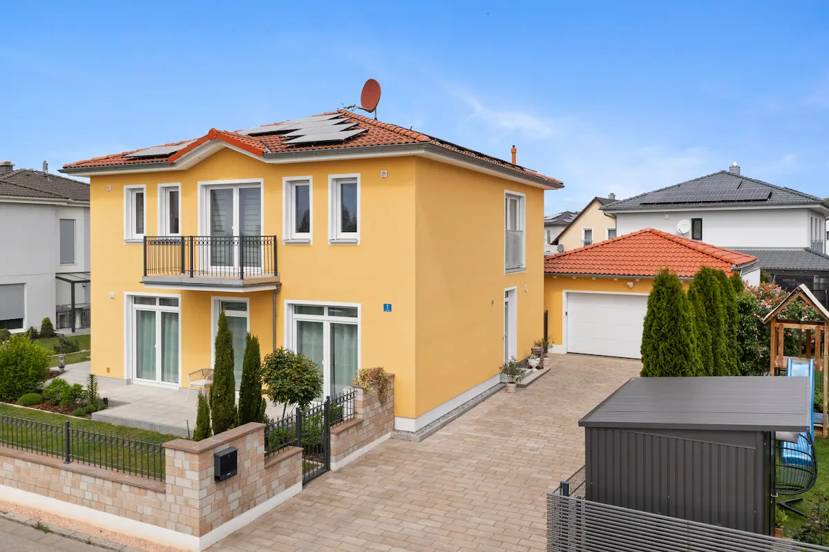 Gelbes modernes Einfamilienhaus mit Solarpanelen auf dem Dach und Vorgarten hinter einem Ziegelzaun.