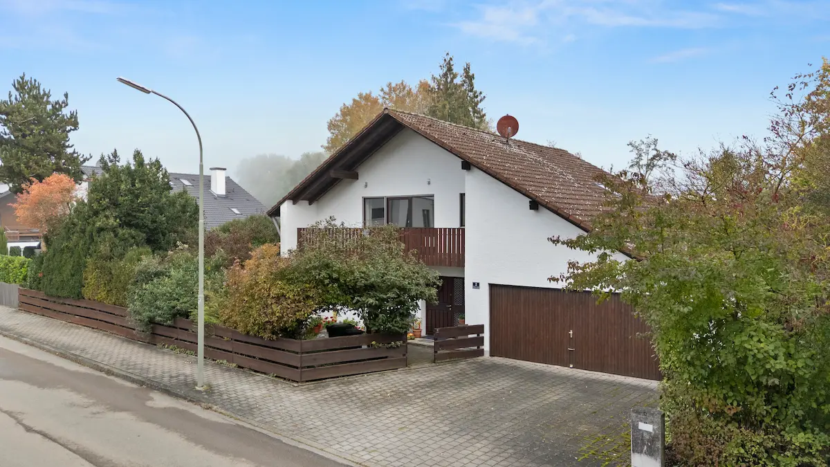 Einfamilienhaus mit weißer Fassade, braunem Holzzaun, Garage und herbstlich bunt belaubten Büschen.