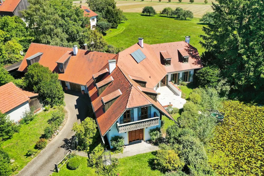 Luftaufnahme eines Hauses mit roten Ziegeldächern, umgeben von Bäumen, Wiesen und einem Teich.