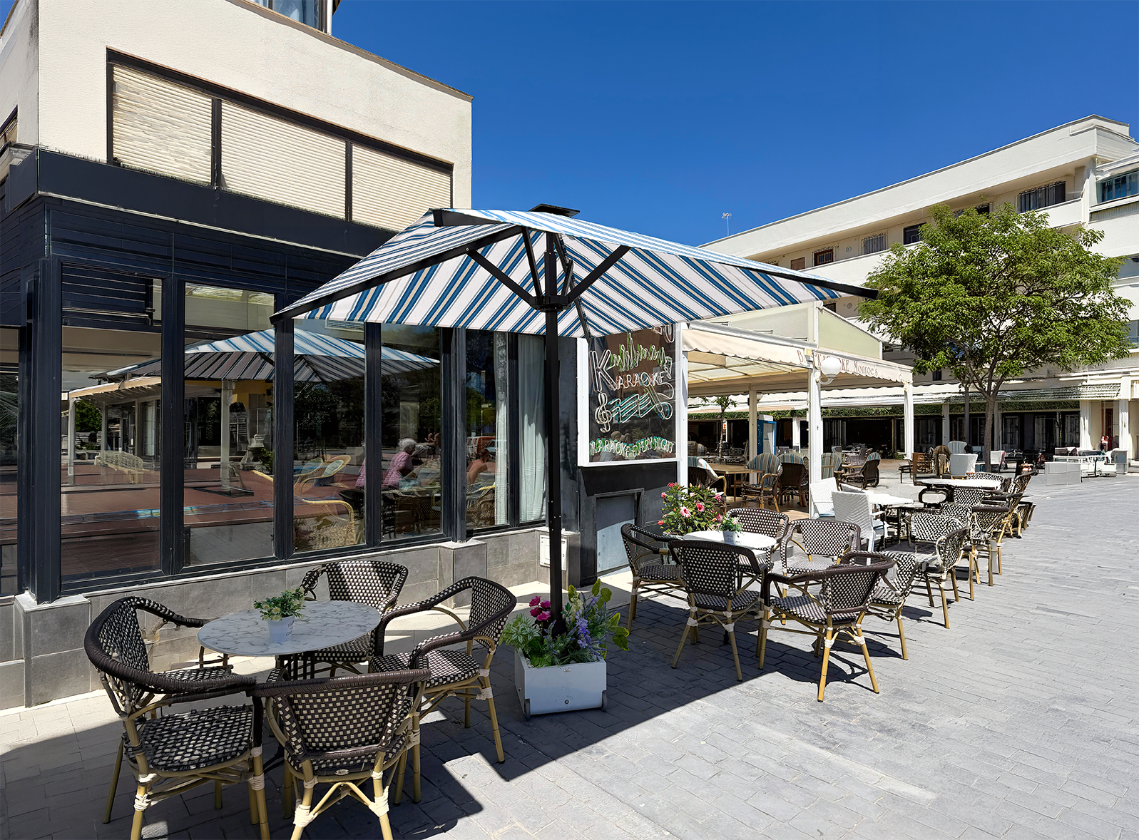 Terraza exterior con mesas y sillas de mimbre bajo sombrillas a rayas frente a un restaurante.